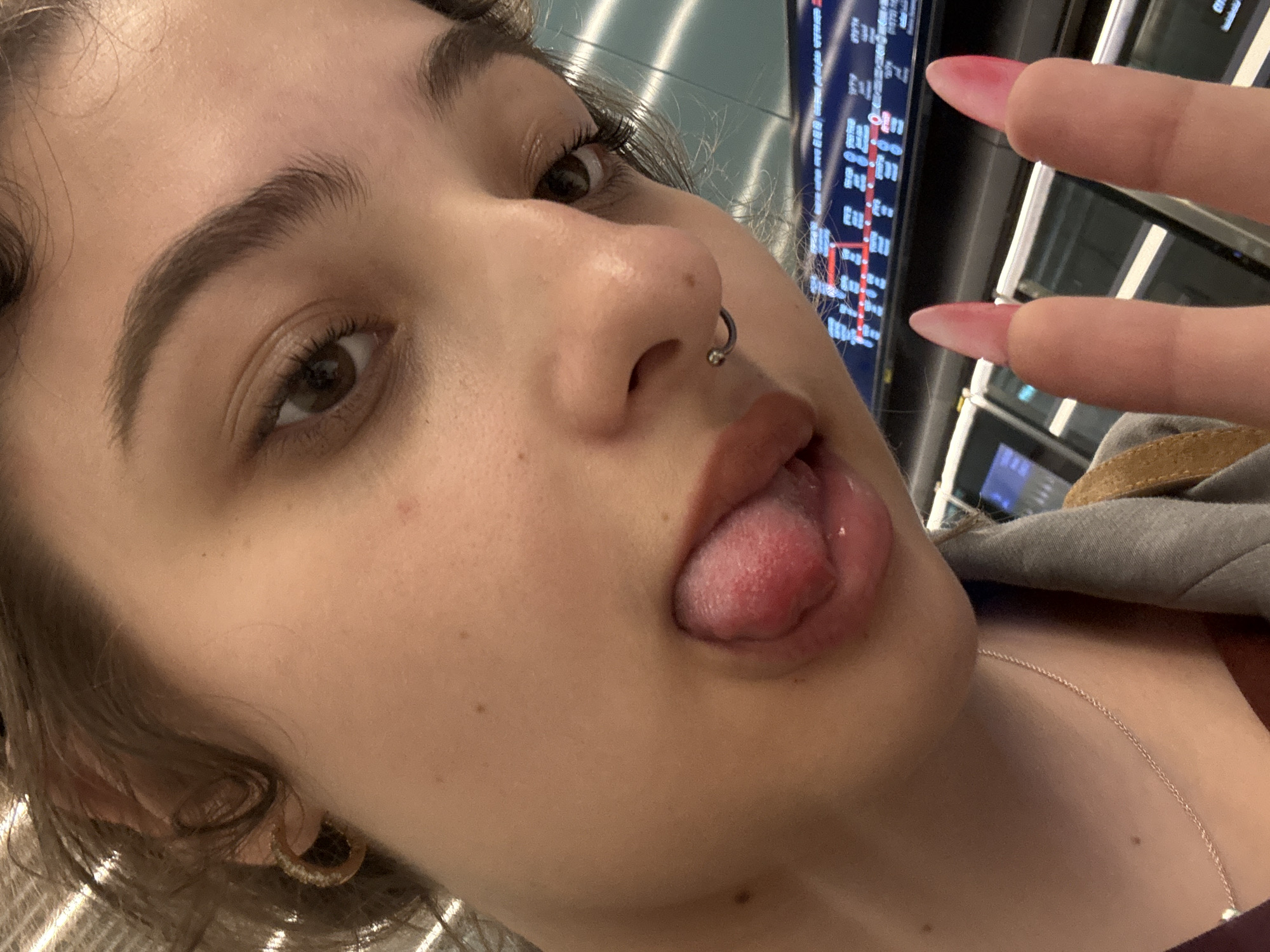 A close-up, low-angle shot depicts a young woman with light brown skin and brown eyes. She has a nose piercing, an earring, and is wearing a thin necklace. Her mouth is open with her tongue protruding. Her fingers with long, pink nails are partially visible at the top of the frame. The background includes a blurred display board with text, likely an arrival/departure board, suggesting a transportation hub. The environment appears to be indoors, possibly an airport or train station. There is no indication of the city or country where this image was taken.