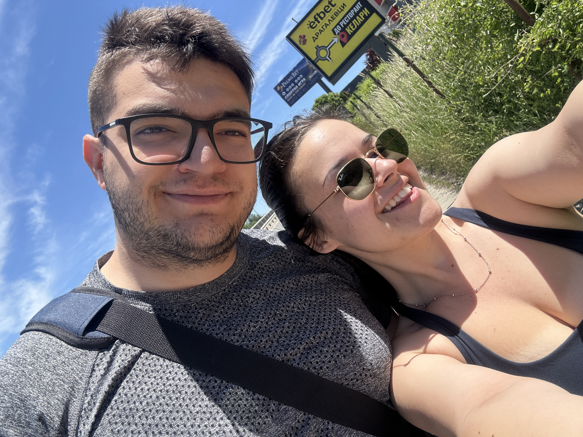 A selfie captures two people in daylight. The person on the left is a fair-skinned man with short, dark hair and a light beard. He wears black-rimmed glasses and a gray, textured shirt with a dark strap diagonally across his body. The person on the right is a fair-skinned woman with her dark hair pulled back, wearing sunglasses and a black top with a delicate necklace. Both are smiling. Behind them are green plants and street signs in Bulgarian, with advertisements for "efbet" and "Palms Bet." The picture was taken in Sofia, Bulgaria.