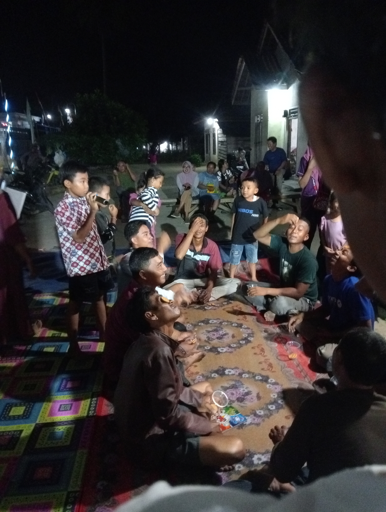 In Rasuan Baru, Indonesia, a group of people are gathered on a patterned mat at night. The scene appears to be outdoors, possibly in a residential area, with buildings and trees visible in the background. The lighting is dim, with artificial lights illuminating the gathering.
Several individuals, including children and adults, are seated or standing. A young boy in a patterned shirt is holding a microphone, suggesting a performance or a social gathering with entertainment. Some people are engaged in conversation, while others are observing. There are plastic cups on the mat, and one person is holding a drink. The atmosphere seems relaxed and social.