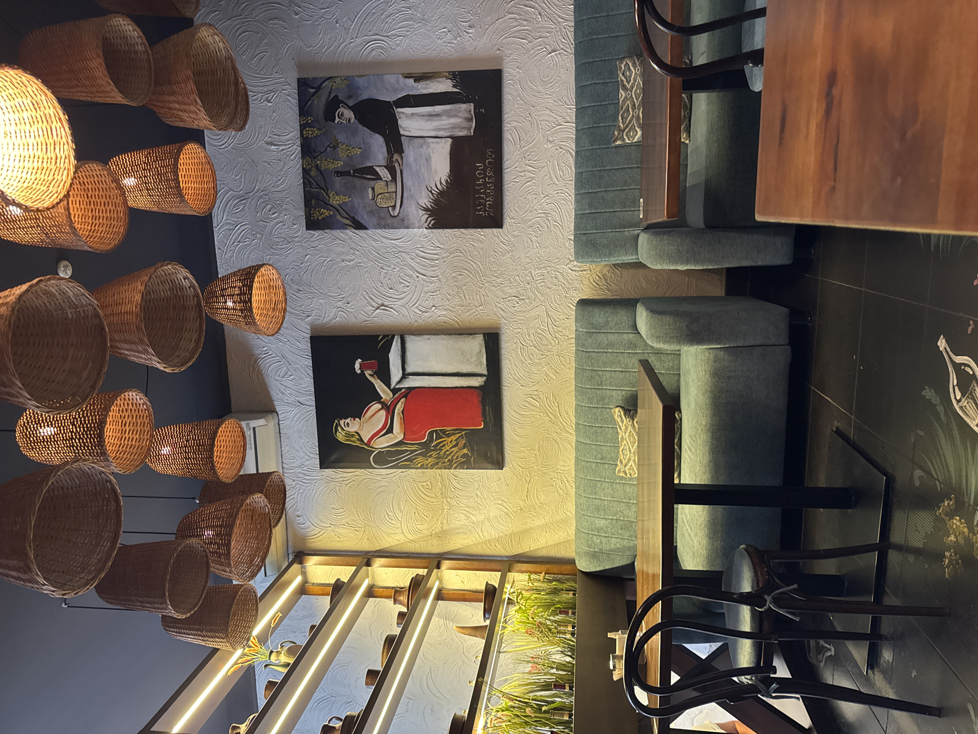 This image depicts the interior of a restaurant in Chernivtsi, Ukraine, characterized by a warm and rustic ambiance. The ceiling features a cluster of woven pendant lights, casting a soft glow on the scene below. On the left, a wall-mounted shelf unit displays a collection of ceramic pottery, some of which are illuminated by integrated LED strip lights.
The walls are adorned with textured wallpaper and two paintings. The painting on the left features a woman in a red dress holding a glass of red liquid, while the one on the right shows a figure in a chef's uniform presenting a tray with a bottle and cheese, below which is text in Georgian script.
Seating arrangements include plush, teal-colored banquettes paired with wooden tables. One table in the foreground is made of dark wood, and a dark wooden chair with a bentwood design is positioned beside it. The floor is tiled in a dark color, with a subtle, light-colored pattern visible. Artificial greenery is arranged along a lower shelf unit, adding a touch of natural element to the decor. The overall impression is a cozy and artistically decorated dining space.