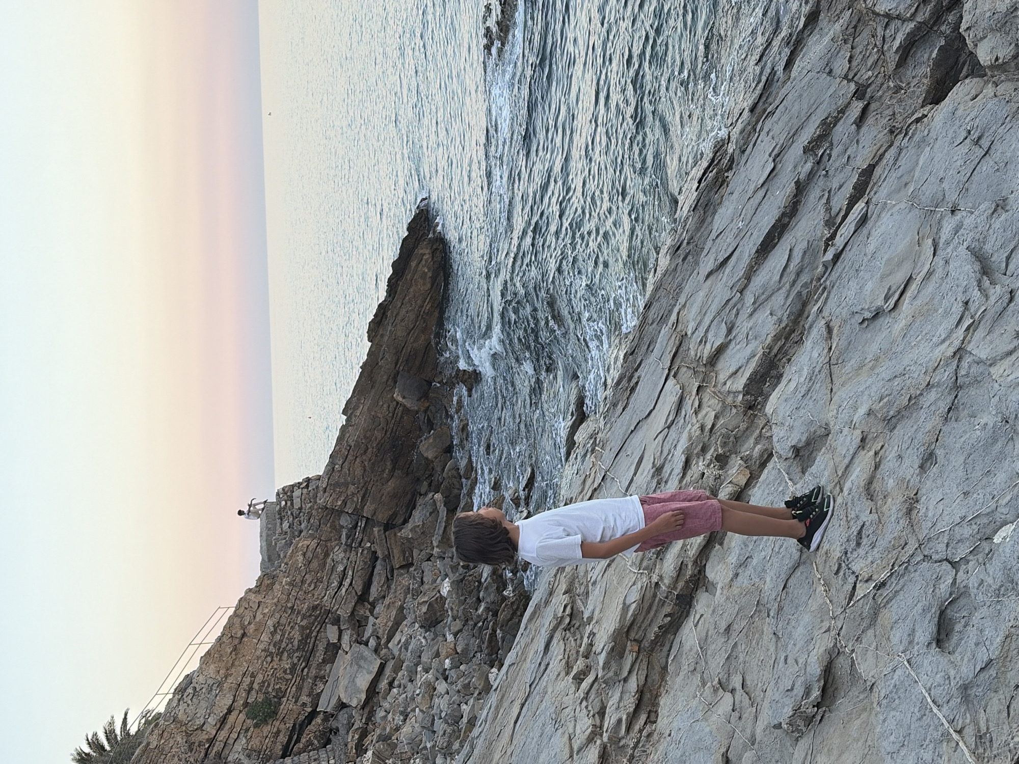 A young boy stands on a rocky coastline, looking up at the sky. He is wearing a white t-shirt and red shorts. The sea is calm and reflects the soft colors of the sunset. The sky is a gradient of pale pink, orange, and yellow, transitioning to a light blue. The rugged terrain suggests a natural, undeveloped area. In the distance, on a rocky outcrop, a small structure resembling a lighthouse or a watchtower is visible, with a person standing near it. There are also some palm trees in the foreground, hinting at a Mediterranean climate. The overall impression is one of peaceful contemplation and appreciation of nature's beauty at dusk.