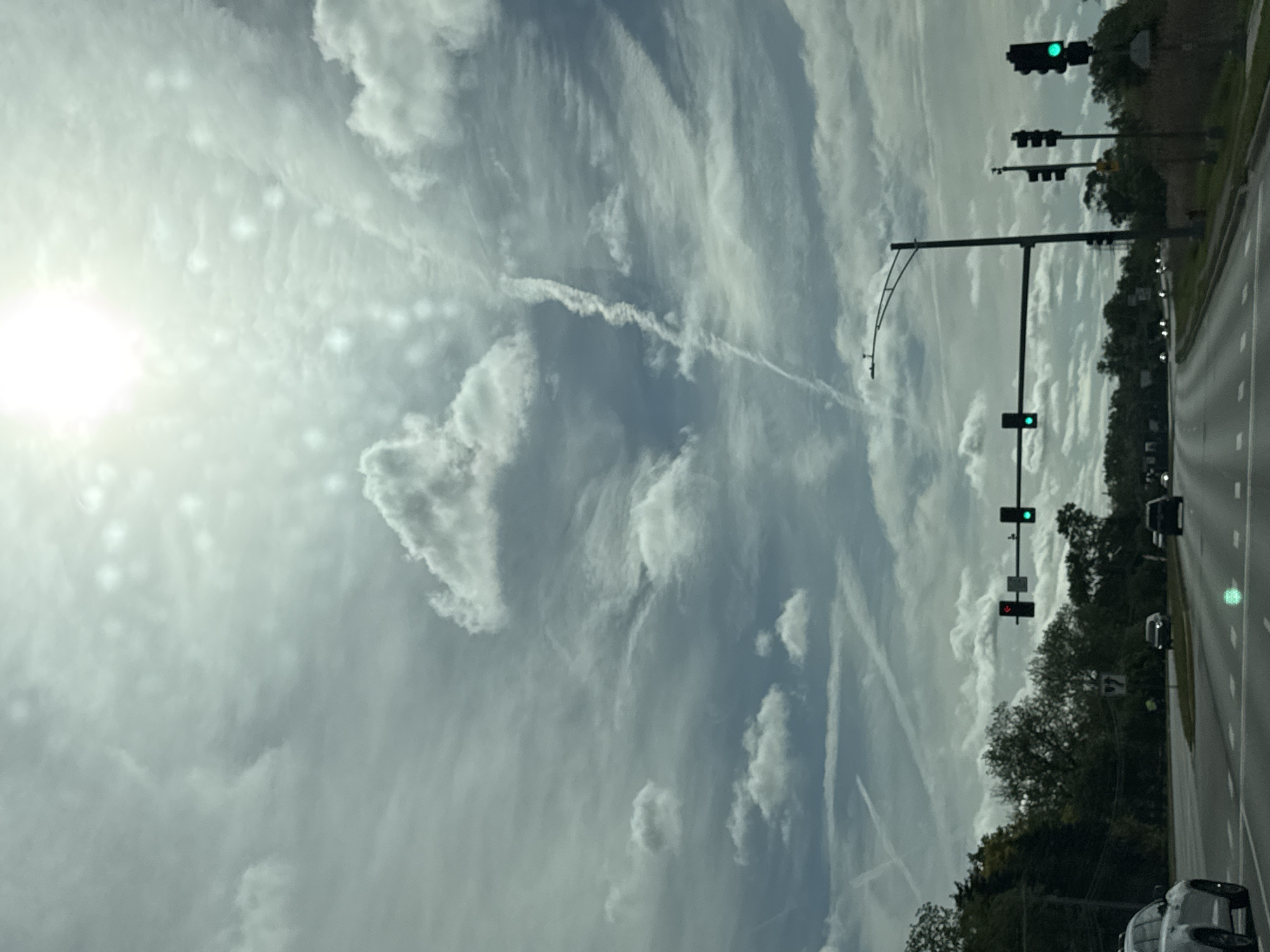 The image captures a scene on a street in Bloomingdale, United States of America. The photograph appears to have been taken from inside a vehicle. A multi-lane road stretches into the distance, bordered by trees and foliage on one side. Traffic lights are visible, some displaying green signals. A few vehicles can be seen on the road. The sky is filled with clouds, and the sun shines brightly in the upper left corner. There are contrails crisscrossing the sky.