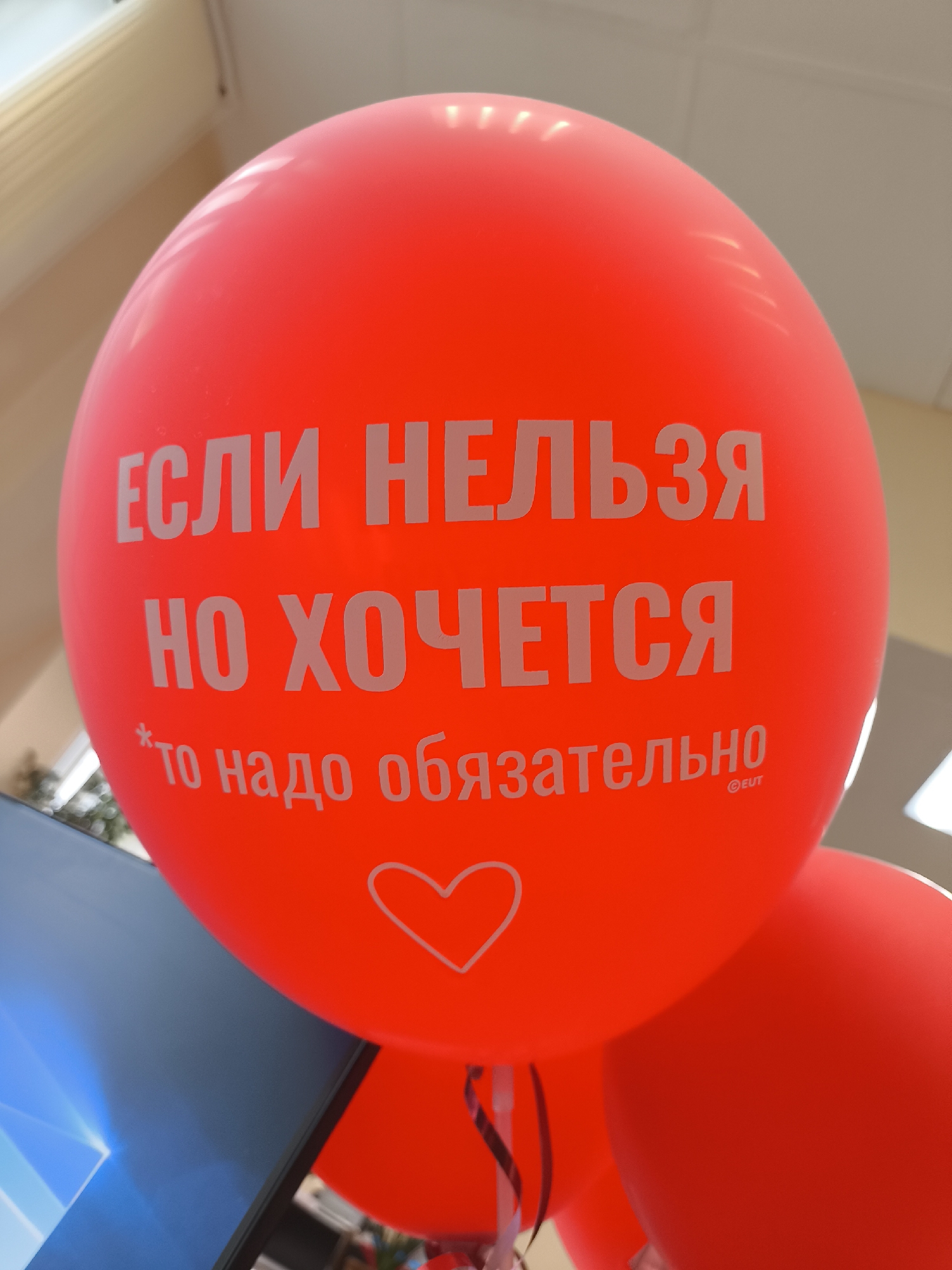 A vibrant red balloon dominates the foreground, featuring white text in Cyrillic script. The text reads: "ЕСЛИ НЕЛЬЗЯ НО ХОЧЕТСЯ *то надо обязательно" (If you can't, but you want to, *then you absolutely must). Below the main text, a small white outline of a heart is visible. A tiny "©BUT" is printed near the end of the last line of text.
The balloon's surface appears glossy, reflecting ambient light from above and to the left, indicating an indoor setting with artificial lighting, possibly fluorescent, as suggested by the ceiling structure in the background. Part of a dark-colored computer monitor with a blue-hued screen saver (possibly Windows 10 default background) is visible on the left side, slightly obscured by the balloon. Another, partially visible red balloon is situated to the lower right. The overall background is out of focus, showing light-colored walls or ceiling tiles and some indistinct elements that could be a window or a plant. The scene suggests a casual environment, possibly an office, retail space, or an event display. There are no people visible, and no specific activity is taking place other than the display of the balloon. The sentiment conveyed by the text is playful and encouraging, perhaps hinting at a celebratory or promotional context.