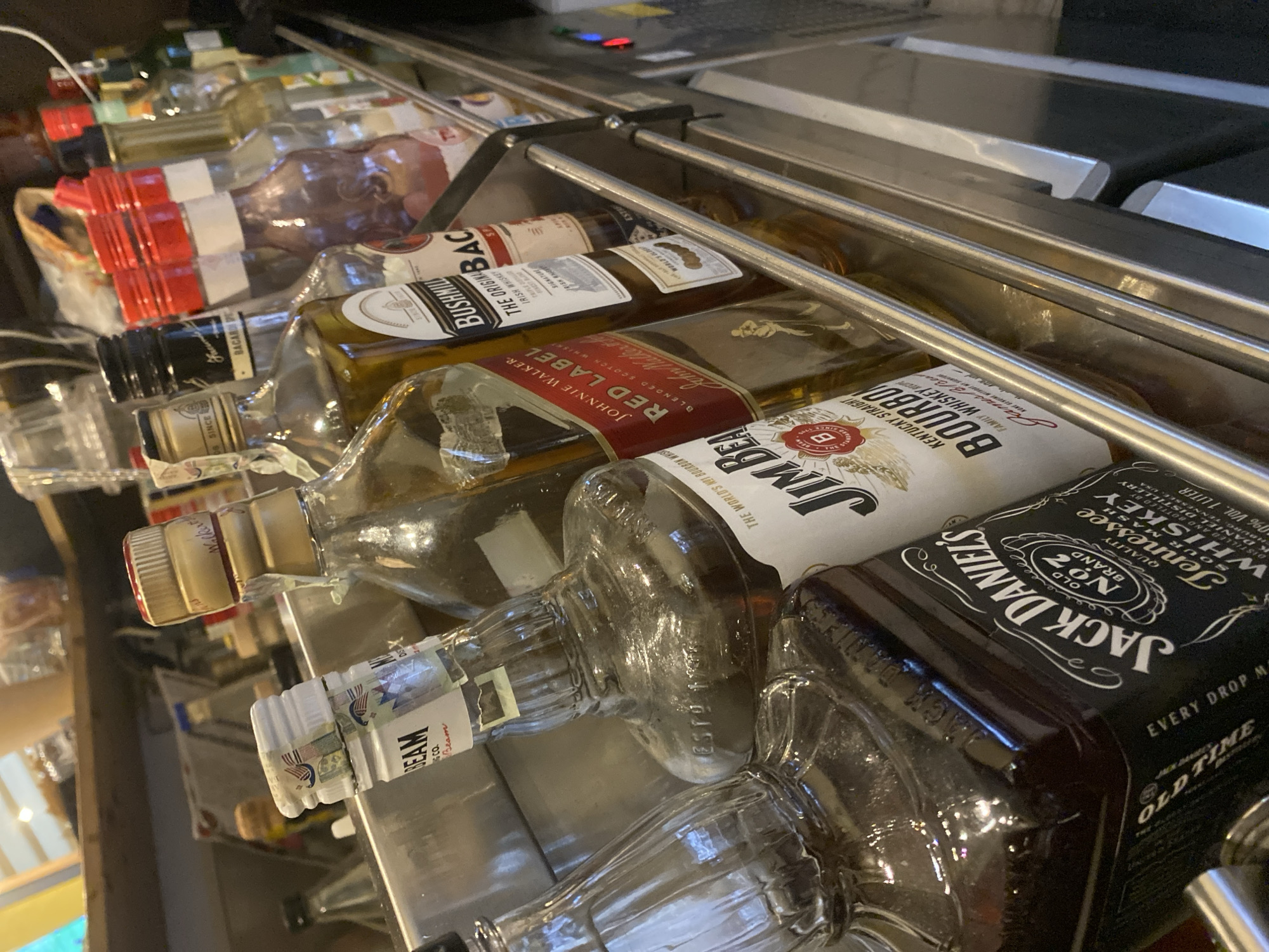 This image shows an assortment of liquor bottles lying on their sides in a tiered metal rack, seemingly behind a bar. The bottles include Jack Daniel's, Jim Beam, Johnnie Walker Red Label, Bushmills, and Bacardi. The setting appears to be a bar or a similar establishment. This image does not specify a location in Varna, Bulgaria.