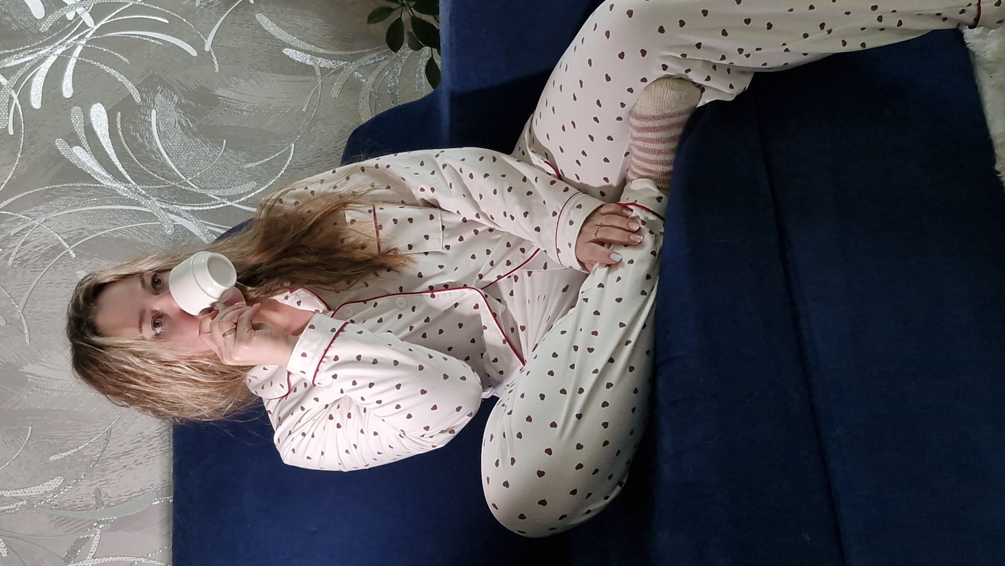 A woman is sitting on a dark blue couch, wearing heart-patterned pajamas and striped socks, and drinking from a white cup. The background features a wall with a silver swirled design and a plant. The location is not specified or identifiable.