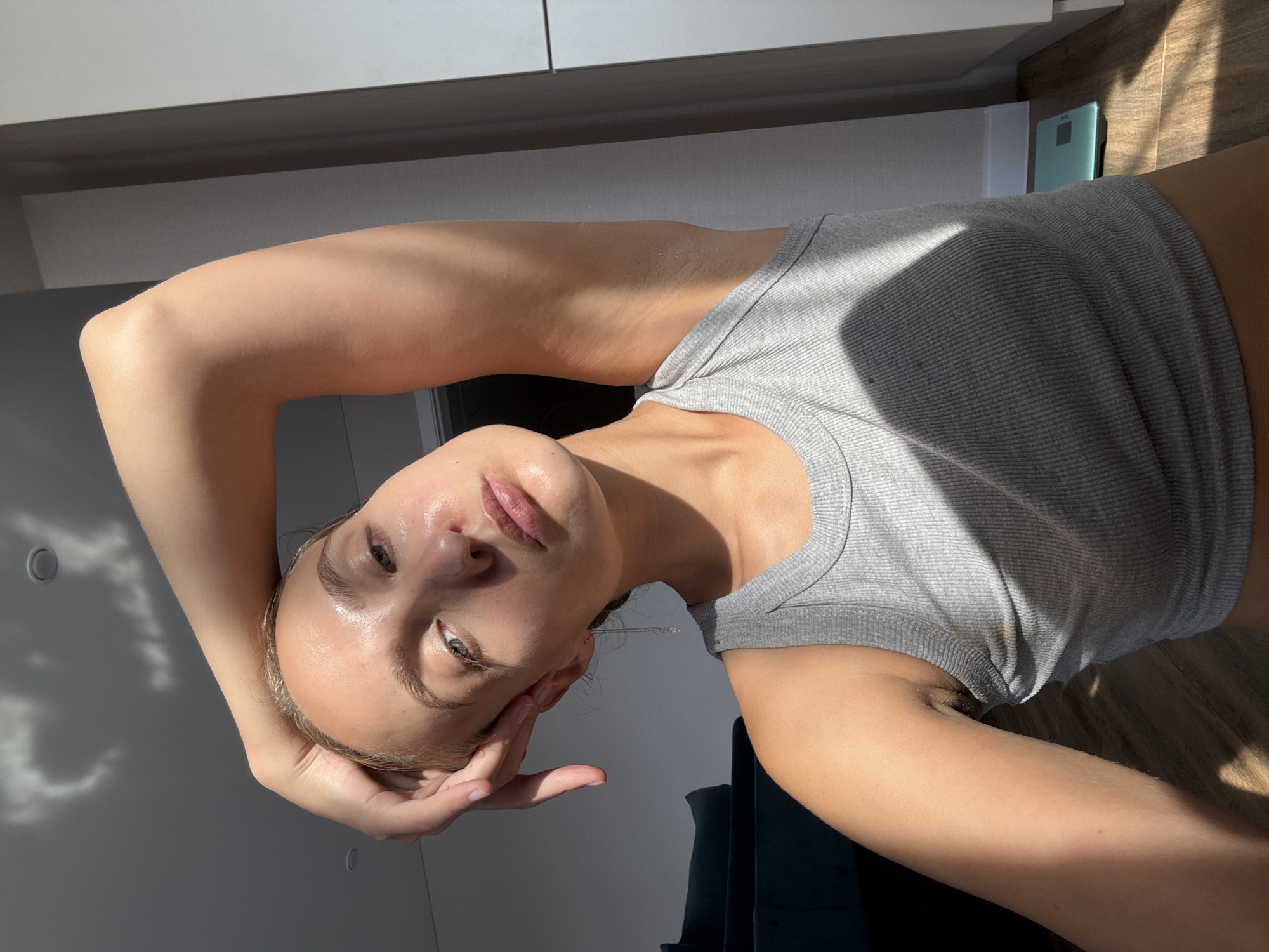 A young woman with blonde hair pulled back is positioned in the center of the frame. Her left arm is raised, with her hand resting on her head, creating a shadow that falls across her face and chest. She is wearing a grey ribbed tank top. Sunlight streams in from the left, casting distinct shadows on her skin, clothing, and the floor. The background shows a dark blue sofa on the left, a white wall and door frame on the right, and wooden flooring. A light blue scale is visible on the floor to the right. The overall scene suggests an indoor environment, possibly a living space, with natural light playing a significant role.