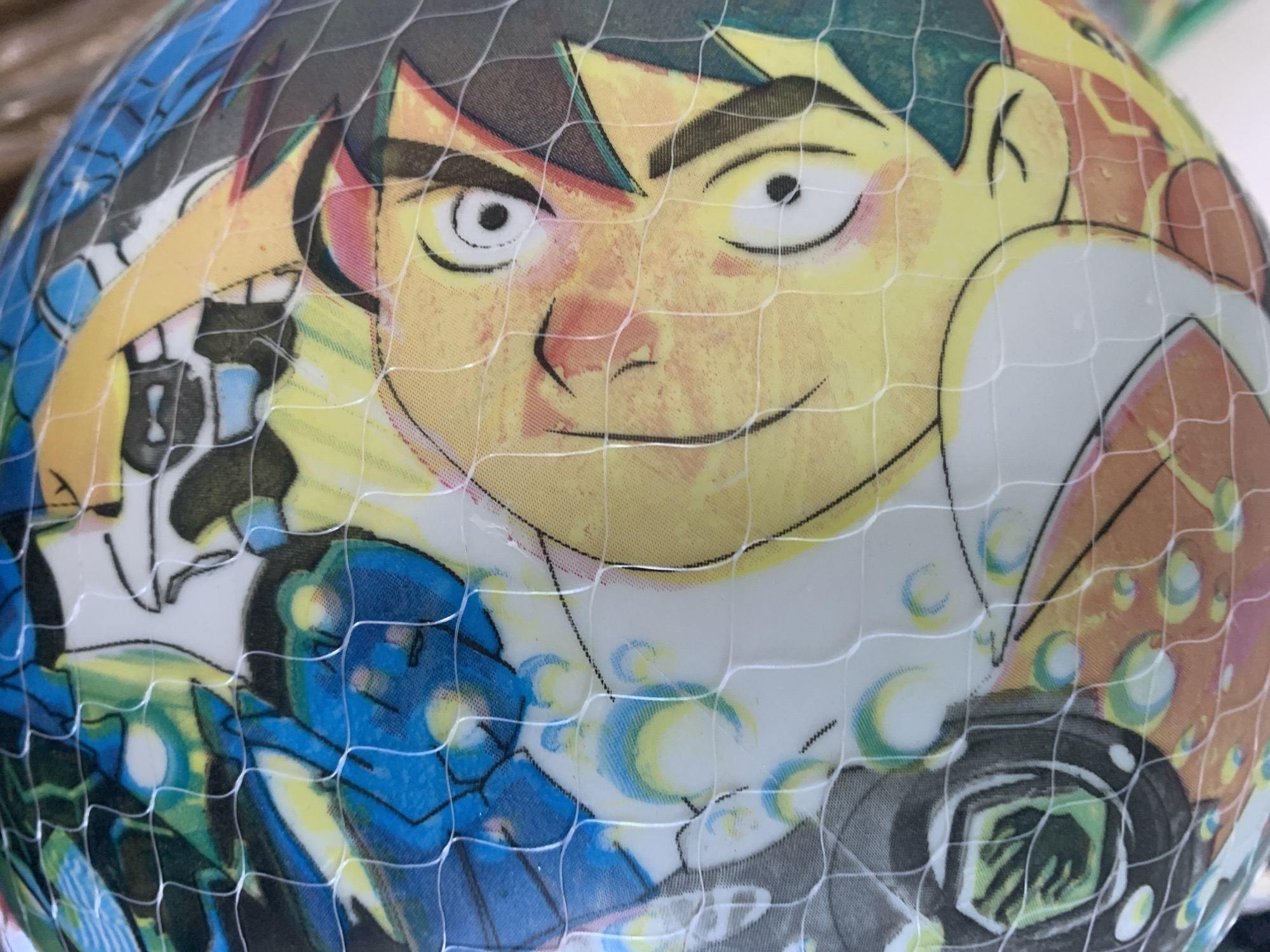 The image is a close-up, angled shot of a spherical object, possibly a ball, covered with a net. The object's surface features a colorful cartoon-style illustration. Prominently displayed is a character with a large, yellow face, wide-open eyes with black pupils, and rosy cheeks. The character is winking with one eye and has black hair with spiky yellow accents. The illustration also includes elements of blue and green, with abstract shapes and lines, possibly representing a background or other characters. A white net with a diamond pattern is draped over the object, obscuring parts of the illustration and creating a textured overlay. The lighting appears to be diffuse, with some highlights and shadows visible on the curved surface of the object and within the net. There is no identifiable location in the city of Unknown, Unknown.