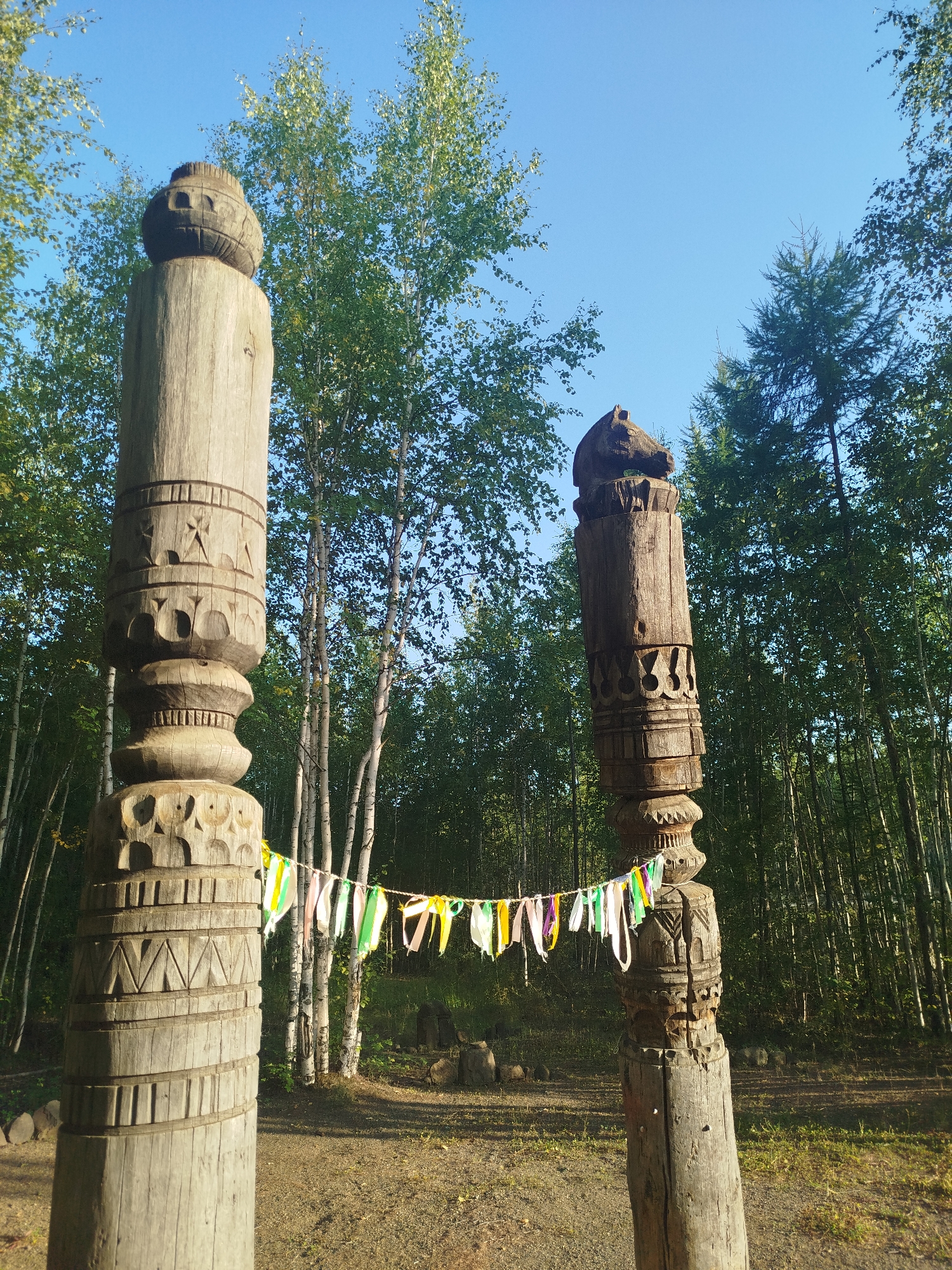 The image shows two large, intricately carved wooden totems standing outdoors in a forested area. A string of colorful prayer flags hangs between them. The totems are made of weathered wood and feature various patterns and carvings, including what appear to be faces or animal heads at their tops. The background consists of a dense forest with tall, slender trees, likely birches, and a clear blue sky. The ground is covered with dry grass and dirt, with some scattered rocks. This scene is likely located in the city of Mirny, Russia, given the context provided. There are no people visible in the image.