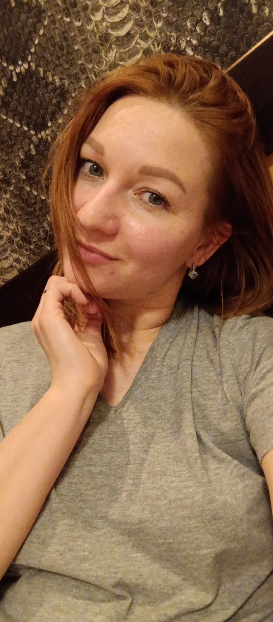 A woman with reddish-brown hair and light blue-green eyes looks directly at the camera in what appears to be an indoor selfie. Her hair is styled around her face, with some strands falling over the left side of her chin. She has a scattering of freckles on her nose and cheeks and a gentle, inviting smile. Her left hand is raised, with her fingers curled and resting against her jawline. She is wearing a simple light grey V-neck t-shirt. A small, geometric-shaped earring is visible on her right ear. The background features a dark, textured surface, possibly a decorative wall covering or upholstery, with a pattern that resembles iridescent scales or overlapping shapes. The lighting suggests an indoor setting, possibly in the evening or at night. No visible text is present in the image.