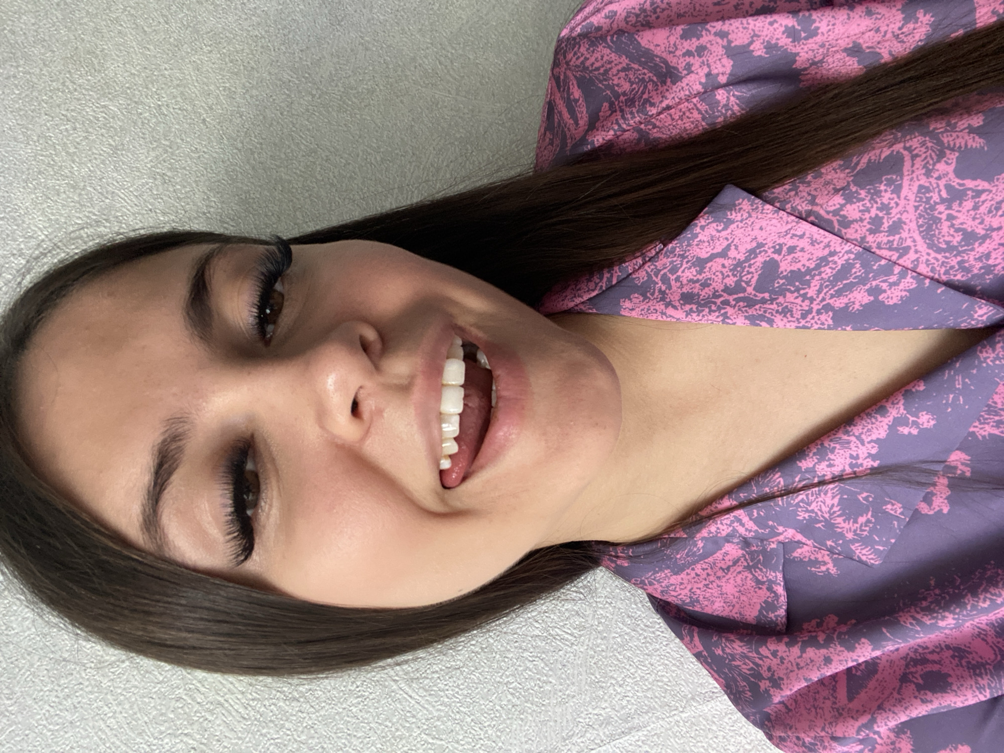 A young woman with long, dark brown hair is smiling broadly and looking upwards. She is wearing dark false eyelashes. She has light skin and is wearing a patterned pink and purple garment. The background is a plain light gray. There is no indication of the location being Chaikovsky, Russia.