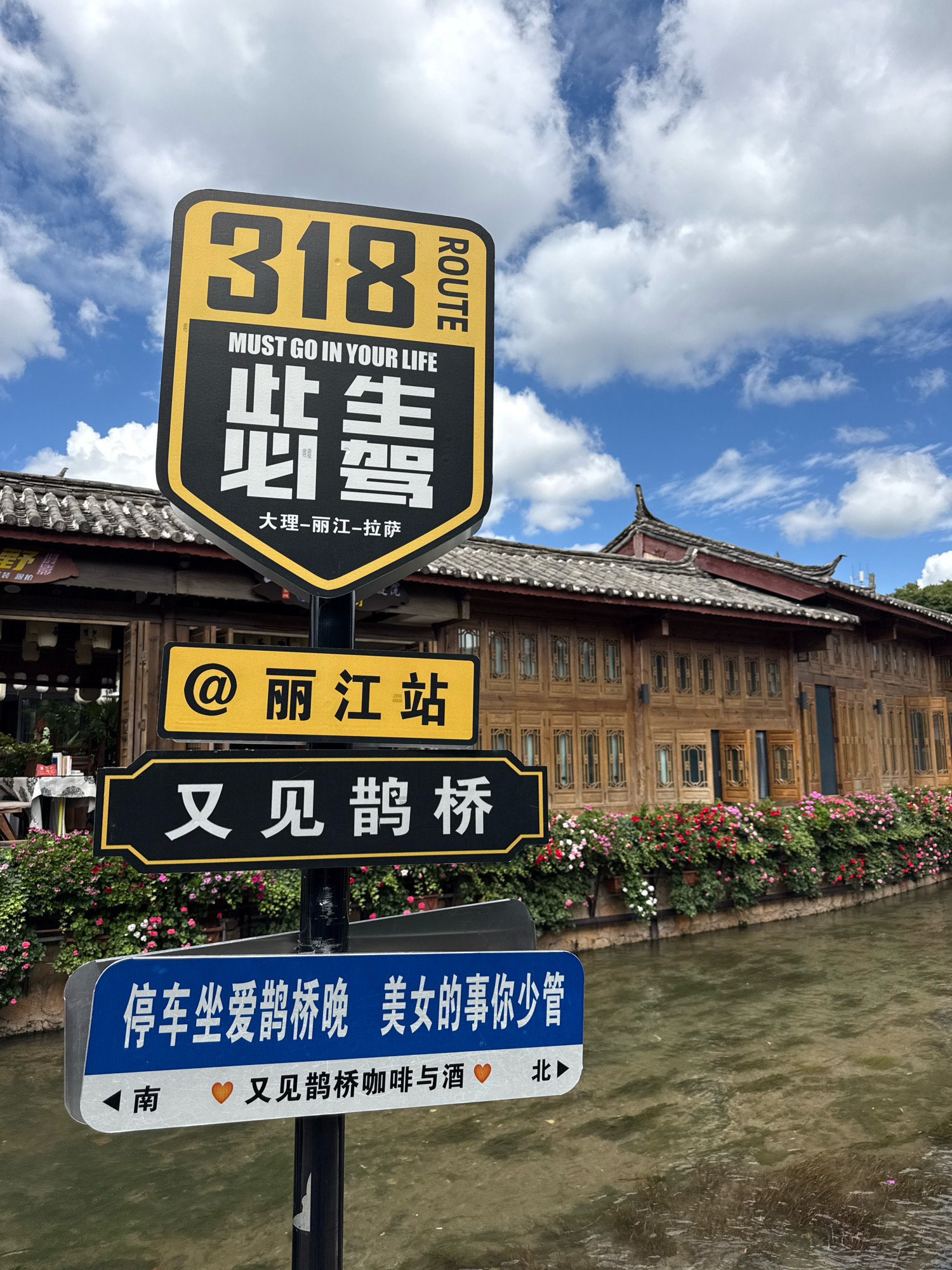 The image shows a street scene in Gucheng District, China, with a canal in the foreground and traditional wooden architecture in the background. Several signs are prominently displayed on a pole. The topmost sign is a black and yellow shield-shaped sign with the number "318" and the text "ROUTE MUST GO IN YOUR LIFE" in English, followed by Chinese characters. Below this, a yellow rectangular sign with a red "@" symbol and Chinese characters reads "Lijiang Station". Another black rectangular sign with white Chinese characters reads "See Goose Bridge Again". At the bottom, a blue and white rectangular sign has Chinese text and arrows indicating directions, with small orange heart icons. The canal is lined with a wall adorned with abundant red and pink flowers. The building in the background features intricate wooden latticework windows and a traditional tiled roof. The sky above is a mix of blue with scattered white clouds. There are no people visible in the frame.