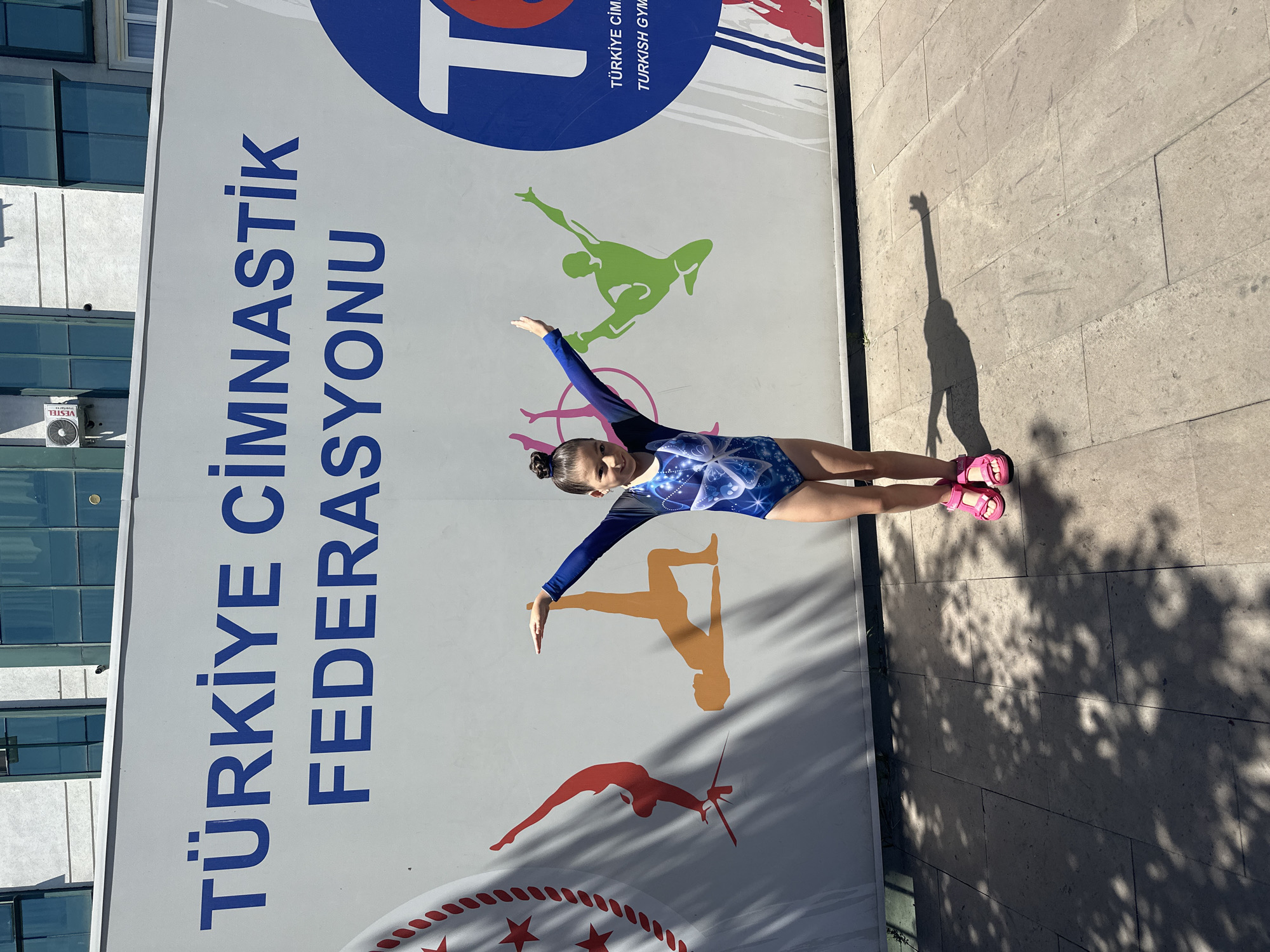 A young girl in a blue and white gymnastics leotard with butterfly patterns and pink sandals poses in front of a large mural on a sunny day. The mural features large blue text that reads "TÜRKİYE CİMNASTİK FEDERASYONU" and stylized silhouettes of gymnasts in red, orange, and green. To the right of the girl, a blue circle contains a large white "T" and smaller text that reads "TÜRKİYE CİM" and "TURKISH GYM". The background is a light-colored building with several windows. The ground in front of the mural is paved with square stone tiles, and the scene is partially covered by shadows from nearby trees. The girl is standing with her arms outstretched, mirroring the pose of one of the silhouettes. The location is identified as Sancaktepe, Turkey.