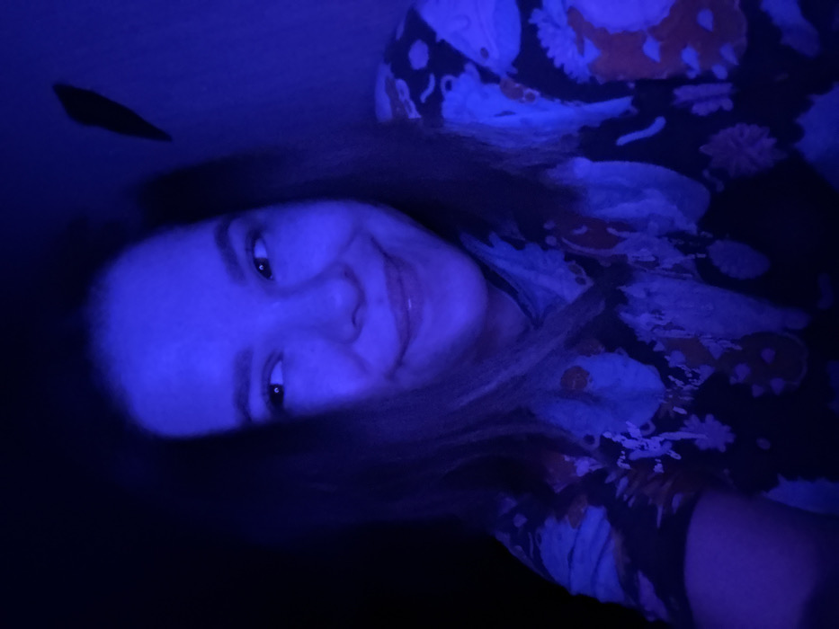 A young adult female is depicted in a close-up, low-light indoor setting in Kazan, Russia. She possesses light-toned skin and dark, medium-length hair. Her face is predominantly illuminated by an intense blue light originating from the right, casting a strong blue hue across her features and visible surroundings. She exhibits a closed-mouth smile and her eyes are directed towards the viewer's upper left. She is wearing a dark-colored top with a discernible, lighter-toned, possibly floral or abstract pattern, heavily tinted by the blue light. The background to the immediate left of the subject is dark and indistinct, suggesting deep shadow. To her right, a light-colored wall is visible, also saturated with blue light. An irregular, dark, elongated shape, consistent with a shadow or a distinct object, is present on the upper right section of this wall.
