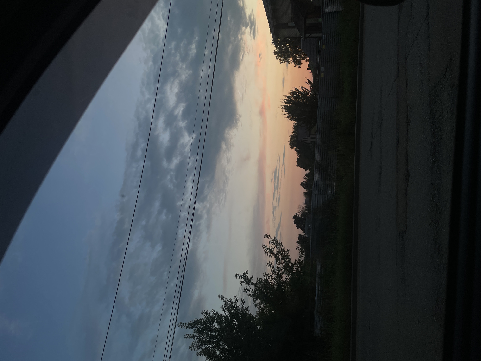 A view from a vehicle looking towards the sky at dusk in Votkinsk, Russia. The sky is a gradient of blue and grey clouds, transitioning to soft pink and orange hues near the horizon. Three power lines are visible stretched across the sky. Below the sky, silhouetted trees and the rooftops of houses are visible. A fence runs horizontally across the mid-ground, separating the trees and houses from the foreground. The foreground consists of a dark asphalt road with visible cracks. The image is framed by dark, indistinct elements on the left and right, suggesting it was taken through a window.