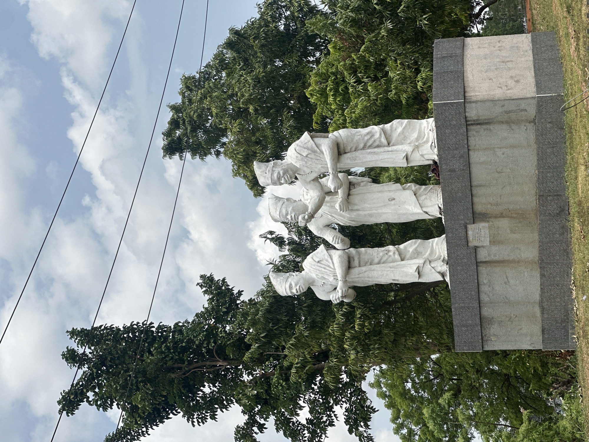 The image shows a statue of three figures on a stone base, likely in an outdoor setting in Ikeja, Nigeria. The figures are dressed in period clothing and appear to be interacting with each other. The statue is surrounded by trees and greenery, under a sky with clouds. Power lines are visible in the upper left corner of the image.