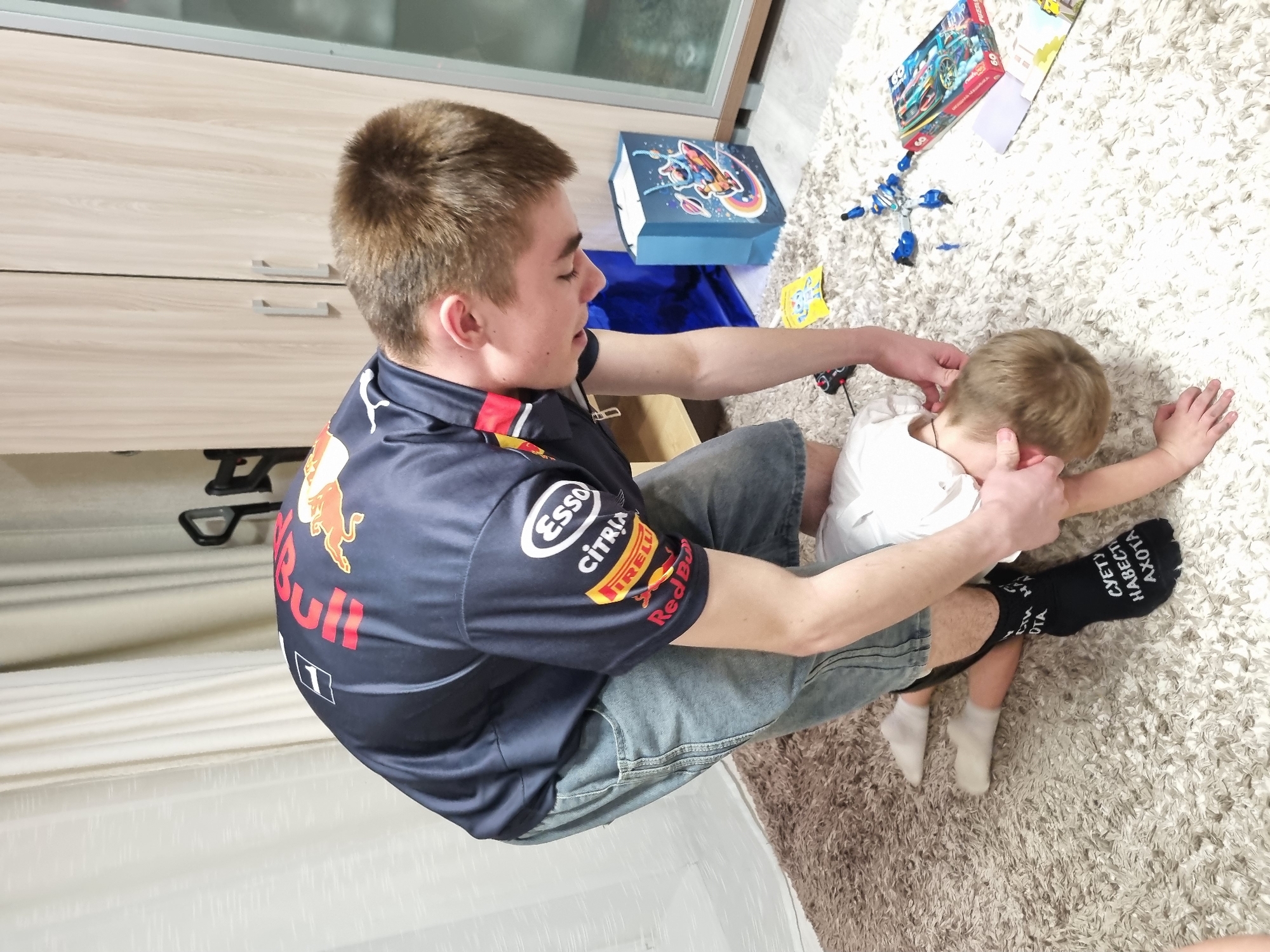 A person in a Red Bull racing shirt is sitting on the floor, attending to a child lying on the carpet. The location is possibly an indoor setting, with a cabinet visible in the background. There are boxes and toys scattered on the carpet near the child. One of the child's socks is black with white text, and the other is white. The exact city of Chaikovsky, Russia, cannot be conclusively determined from the image alone.