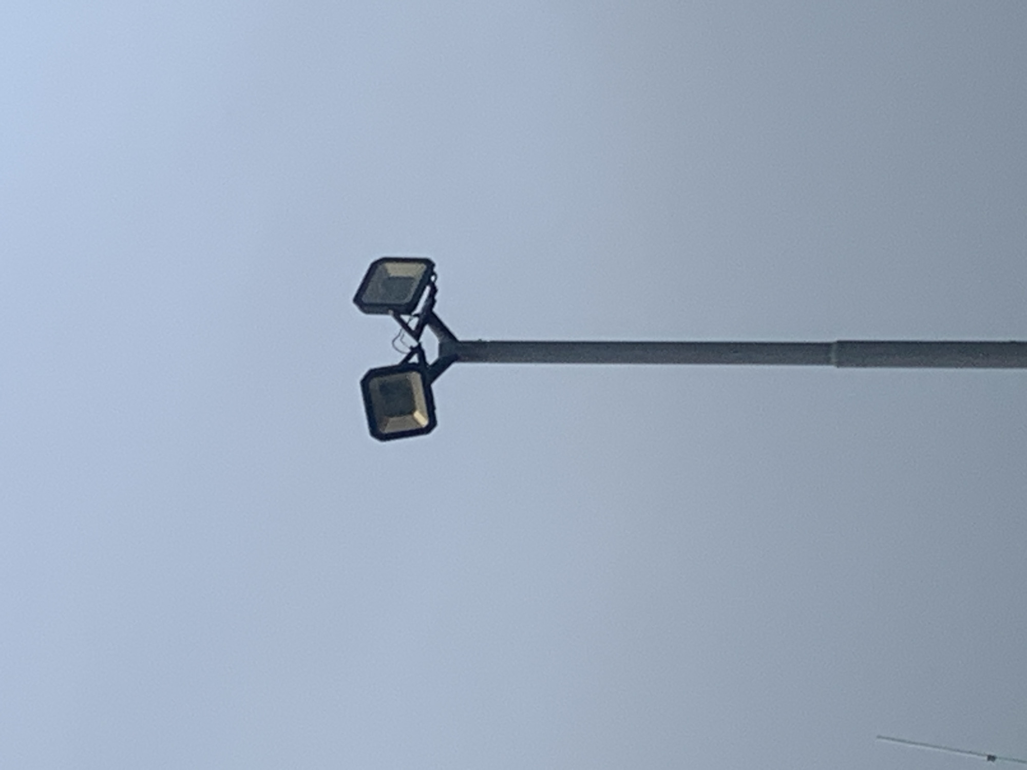 A tall, grey pole supports two square floodlights, one on each side, angled outwards. The floodlights appear to be made of dark metal with illuminated glass fronts. The sky behind is a pale, overcast blue. A thin white pole or antenna is visible in the lower left corner of the frame. The scene is devoid of people or any other significant objects.