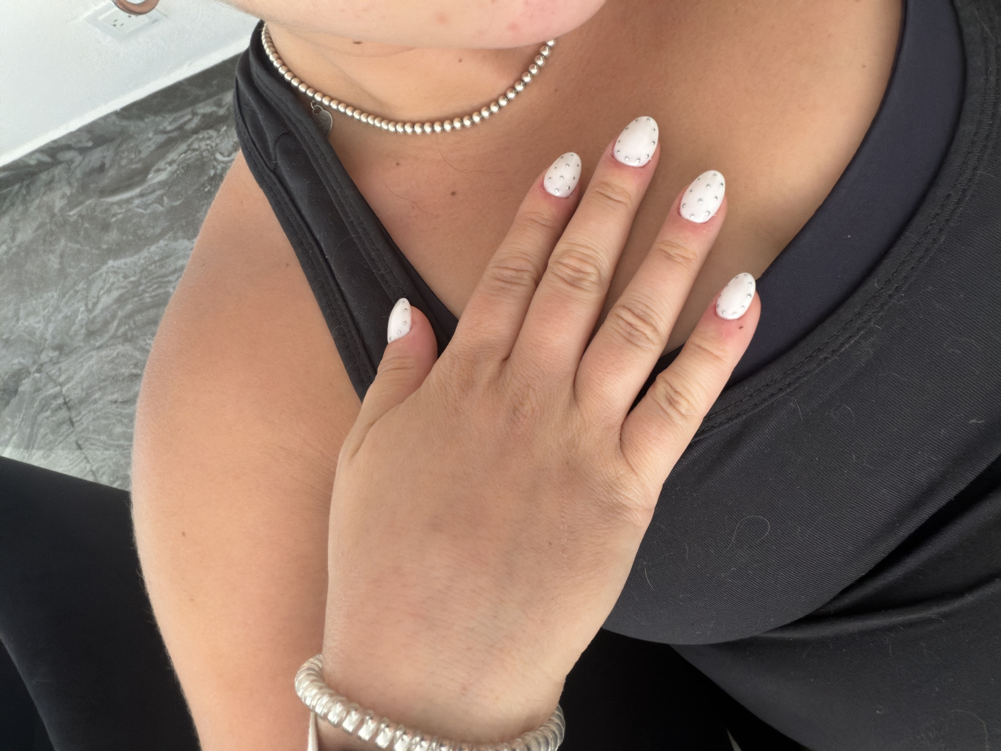A photograph shows a person wearing a black tank top and black pants. The person has a silver bead necklace and a silver bead bracelet. Their hand is resting on their chest, showcasing white nails with small, silver embellishments. The background includes a gray and white marbled surface. The location is unknown and cannot be determined from the given image.
