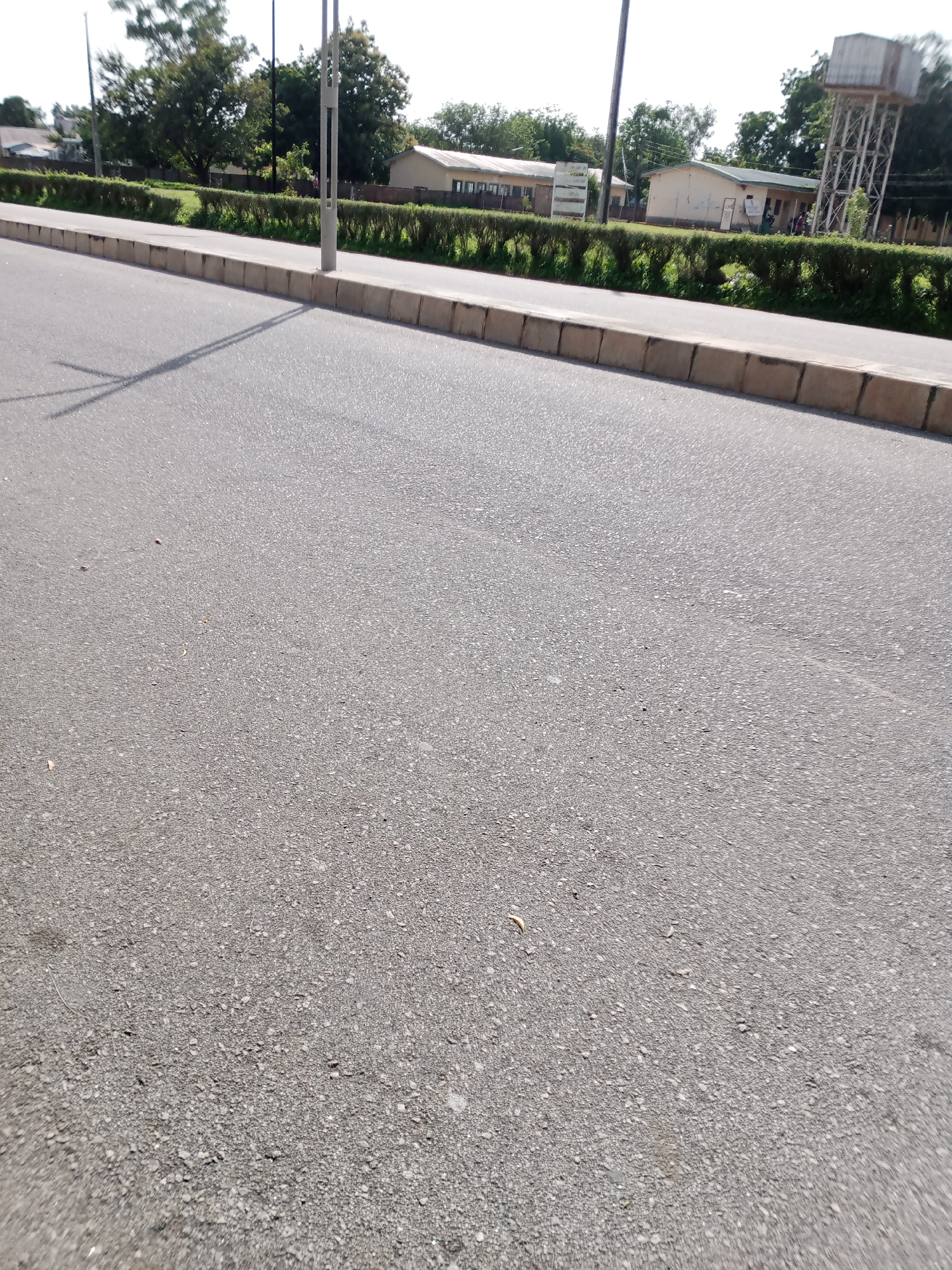 This is a medium-angle, eye-level, outdoor shot of a paved road in Jalingo, Nigeria. The road is gray asphalt with a concrete curb running along the left side. Beyond the curb is a manicured hedge, and behind that are several low-lying, light-colored buildings with tiled roofs, partially obscured by trees. To the far right, a tall, metal water tower with a rectangular tank at the top is visible. The sky is bright and hazy. A street light post with two horizontal lights is positioned on the curb. The shadow of the street light post stretches across the road. There are no people clearly visible in the image. The environment appears to be a public space, possibly a street in front of an institutional or commercial area.