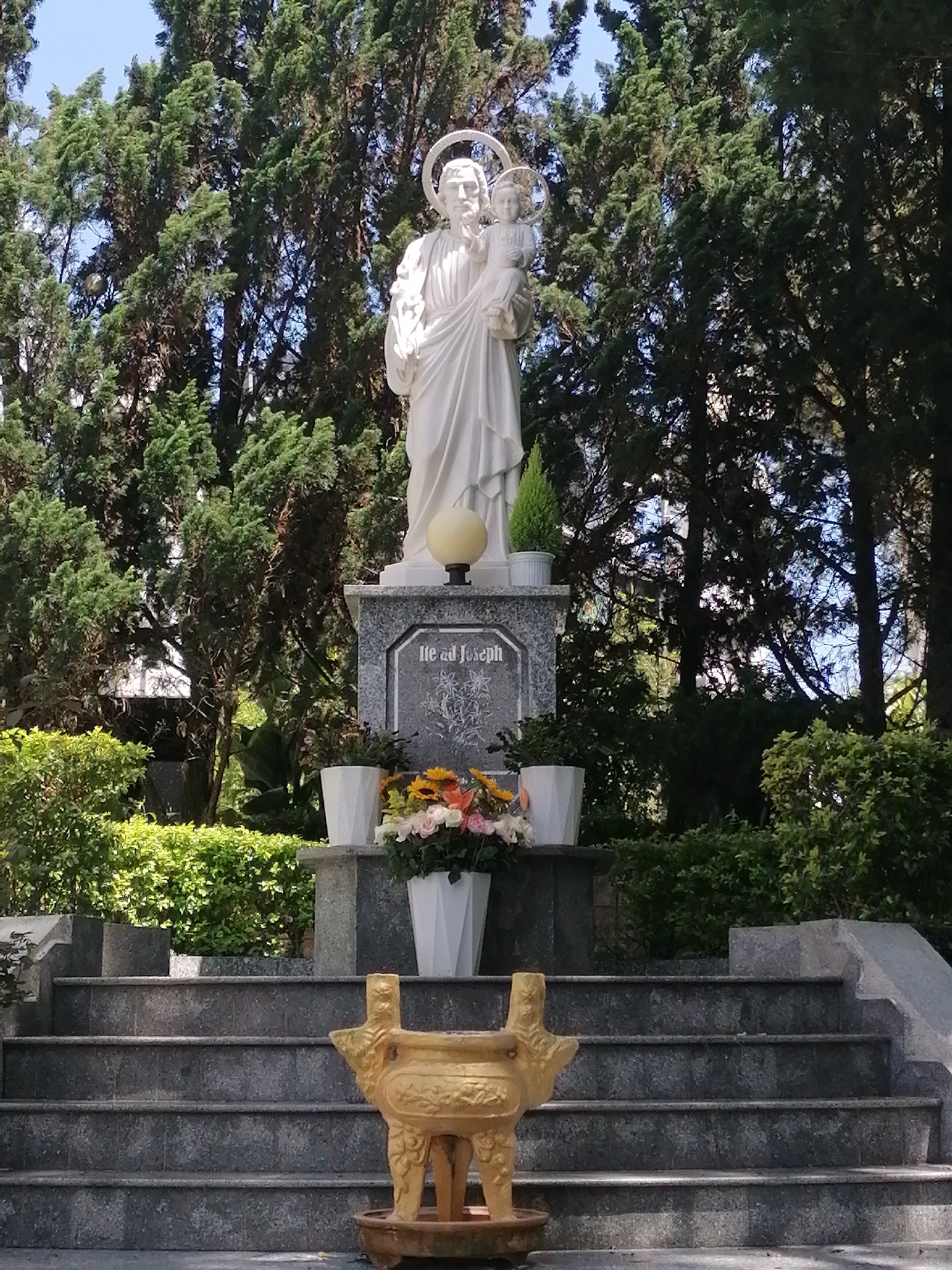The image depicts a statue of St. Joseph holding the child Jesus, standing on a granite base inscribed with "Ile ad Joseph." The statue is surrounded by trees and shrubbery in an outdoor setting. In front of the statue are steps leading up to the base, with flowers and plants arranged on the steps and base. A golden incense burner sits at the base of the steps. The location is Da Lat, Vietnam, based on the typical Vietnamese architecture.