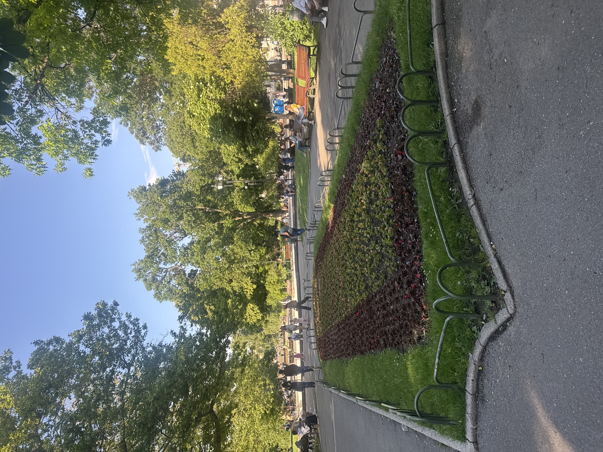 This image captures a vibrant daytime scene in a public park in Sofia, Bulgaria, characterized by lush greenery and a clear blue sky. The photo is taken from a slightly elevated perspective, looking across a paved path towards a central green area lined with trees and garden beds, with more trees framing the top and left of the frame.
Along the right side, a dark asphalt path curves gently, bordered by a concrete curb and a low, decorative green metal fence. Beyond this fence are meticulously maintained garden beds featuring densely planted shrubs with dark red and green foliage, interspersed with patches of bright green grass.
In the middle ground, numerous people are scattered throughout the park. Many are seated on classic wooden park benches, some conversing, others seemingly relaxing. Other individuals are seen walking along the paths or standing amidst the trees. The crowd suggests a popular spot, possibly during a warm day. On one bench, two distinctive blue cylindrical objects (possibly public bins or containers) are visible near people.
The scene is dominated by tall, deciduous trees with rich green leaves, providing ample shade. The sunlight filters through the leaves, creating dappled patterns of light and shadow on the ground. The sky is a bright, cloudless blue, indicating fair weather. The overall atmosphere appears tranquil and inviting, with people enjoying the pleasant outdoor environment. There is no visible text in the image.