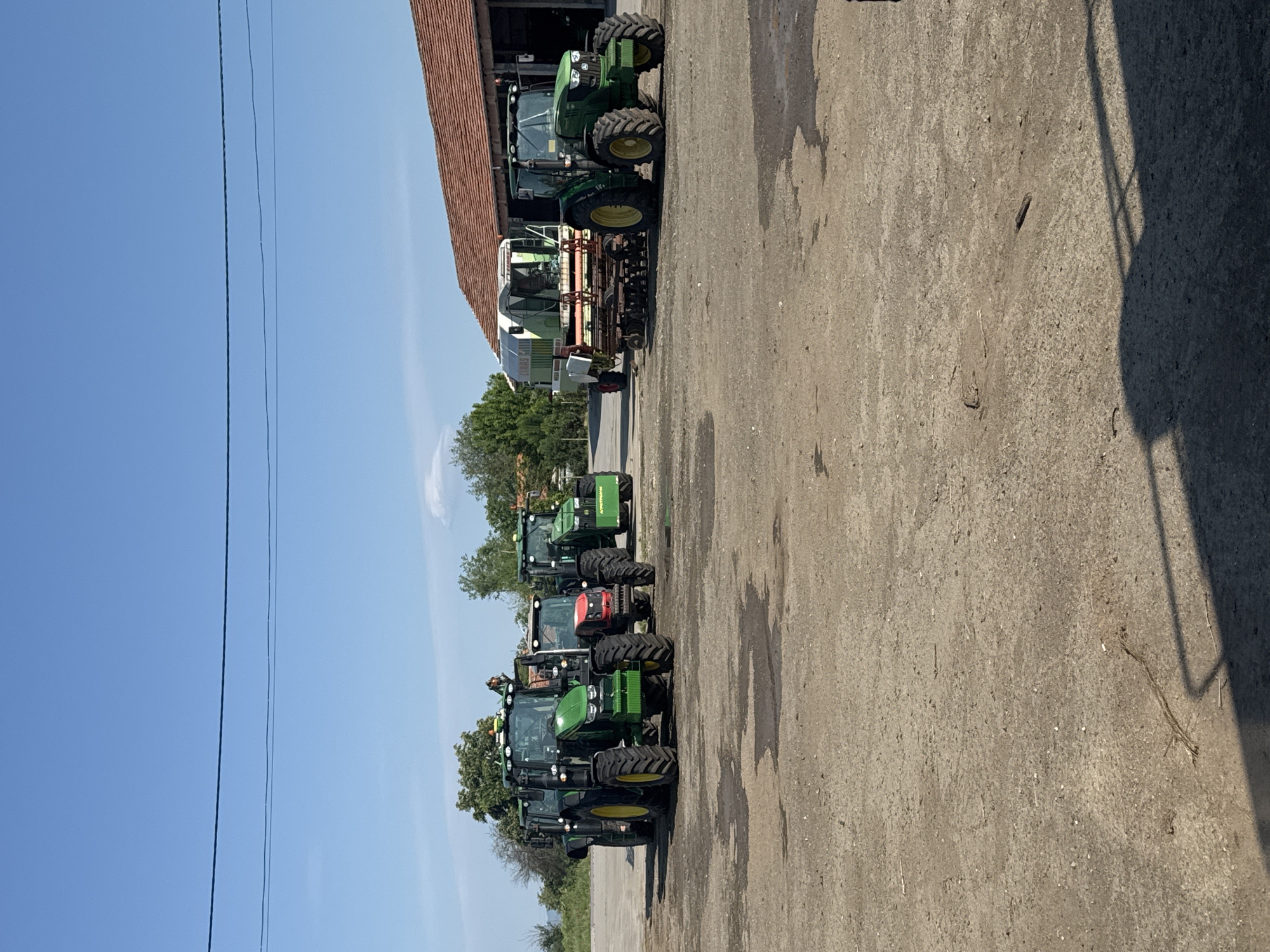 The image shows a row of tractors parked on a concrete surface. The tractors appear to be in Sarnevo, Bulgaria. The vehicles are mostly green and yellow and are parked alongside a building with a red roof. A clear blue sky is visible in the background. There are trees in the background. Power lines are visible in the sky. Shadows are cast across the concrete.
