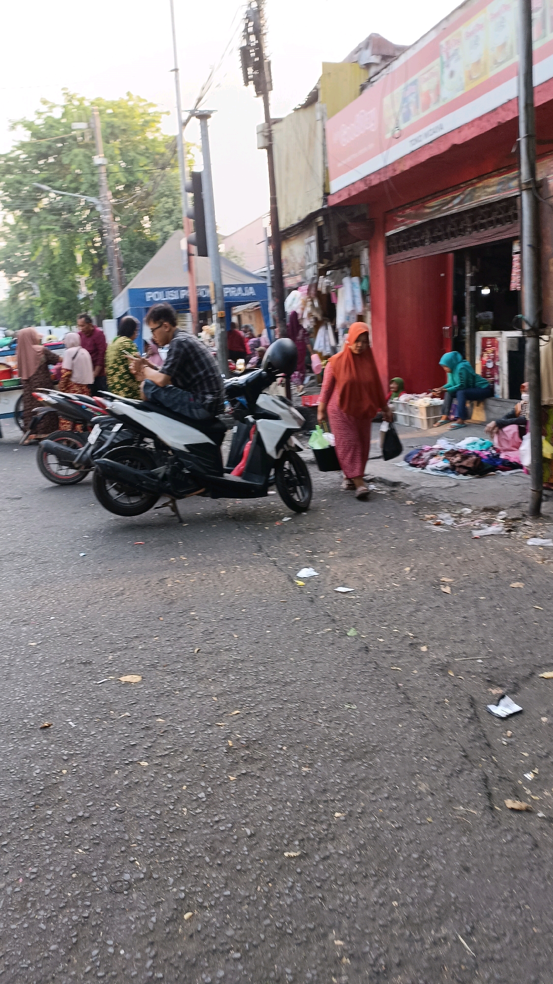 A street scene in Surabaya, Indonesia shows a person on a motorcycle. The motorcycle is white and black, and the rider is wearing a black and white checkered shirt. They are looking down at a smartphone. A woman in an orange headscarf and pink dress walks past, carrying two bags. In the background, there is a building with a red facade and a sign that reads "Cool City". Other people and market stalls are visible. The street is paved with asphalt and has some debris on it. Trees and a traffic light are also present in the background.