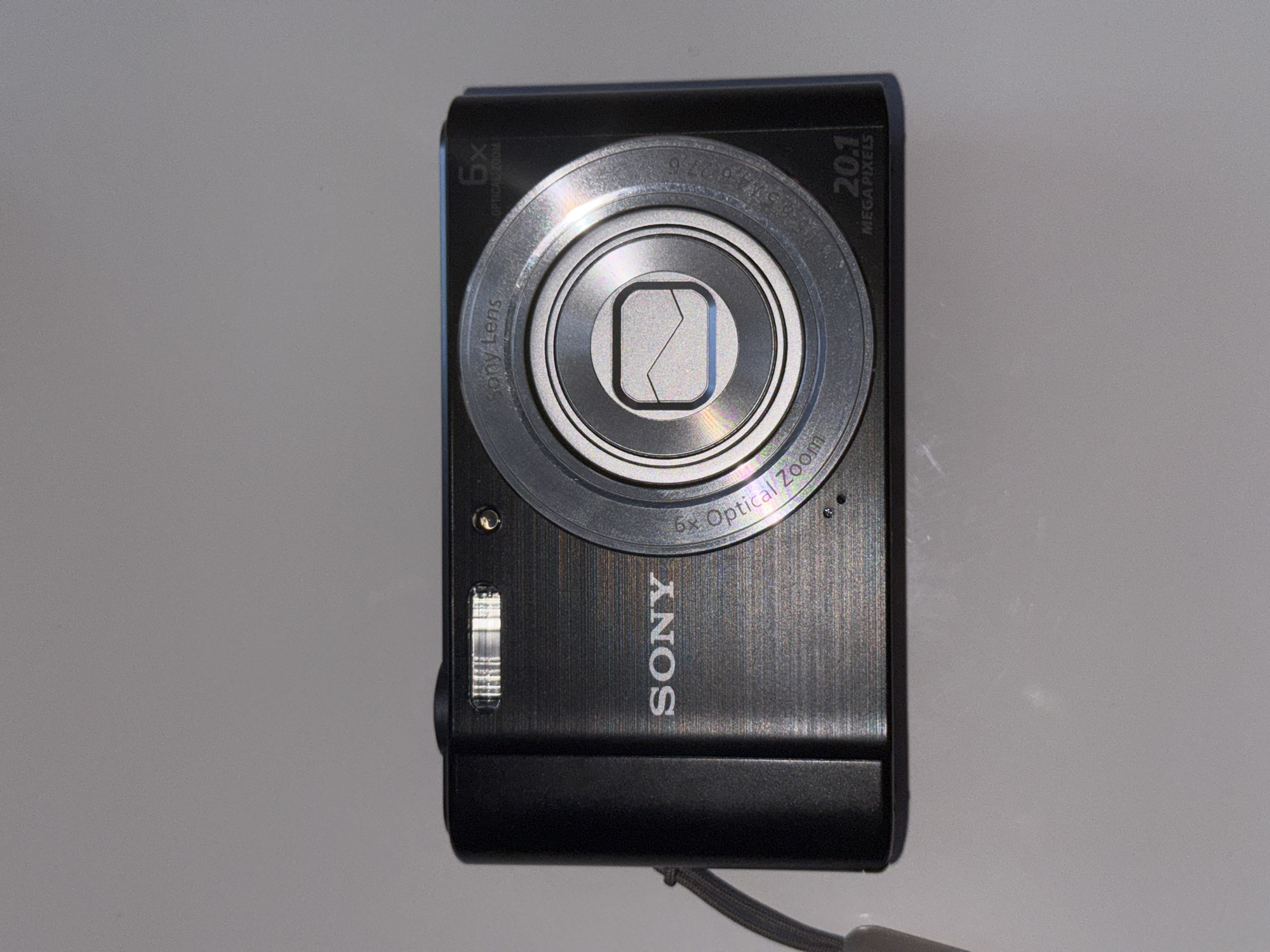 A black Sony digital camera is depicted in a top-down, vertical orientation against a plain, light grey background. The camera's main lens assembly, a prominent silver circle, is centered in the upper portion of the frame. Within this lens assembly, a metallic silver shutter or lens cover with a distinctive geometric, almost abstract 'Z' or 'N' shape, is visible.
The camera body has a brushed finish, while a small silver light or flash is situated on the left side of the camera, next to the lens. A grey wrist strap protrudes from the bottom right corner of the device.
Several details are legible on the camera. The brand name "SONY" is emblazoned vertically below the lens. Around the silver lens ring, text reads "SONY Lens" and "9.70-46.60mm 1:3.3-6.3" at the top, and "5x Optical Zoom" at the bottom right. On the black camera body itself, "6x OPTICAL ZOOM" is visible on the upper left side, and "20.1 MEGAPIXELS" is inscribed on the upper right, indicating its optical zoom capability and sensor resolution.
The lighting is even, suggesting an indoor product shot. The image provides no specific activity, focusing solely on the camera itself. The location context for this item is Lobanovo, Russia.