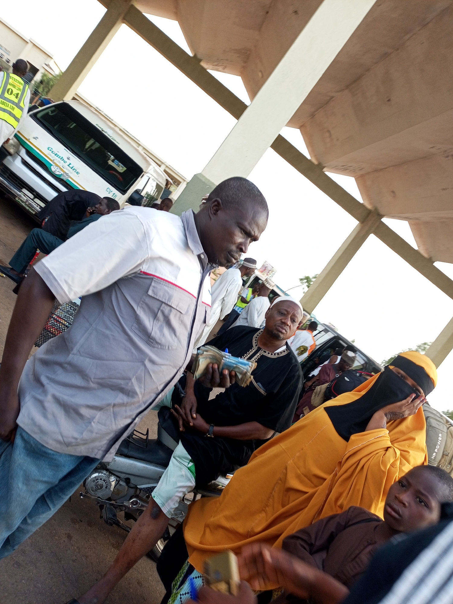 An outdoor scene at what appears to be a bus station or loading area in Gombe, Nigeria, under a beige canopy. In the foreground, a man in a light gray and white short-sleeved shirt with red trim and blue jeans is seated on a motorbike, looking to the right. Beside him, a woman wearing a bright yellow hijab and niqab is seated, with her hand covering her mouth and eyes. To her right, a young boy in a brown garment is looking towards the viewer.
In the background, a white minibus with green and teal stripes is visible, branded "Gombe Line." Several people are gathered around and inside the minibus. A man in a black shirt with white trim is holding a wad of cash. Another individual in a yellow vest with "STATION 04 GARBA LINE" is standing near the front of the minibus. The ground is paved with asphalt. The overall atmosphere suggests a busy transportation hub.