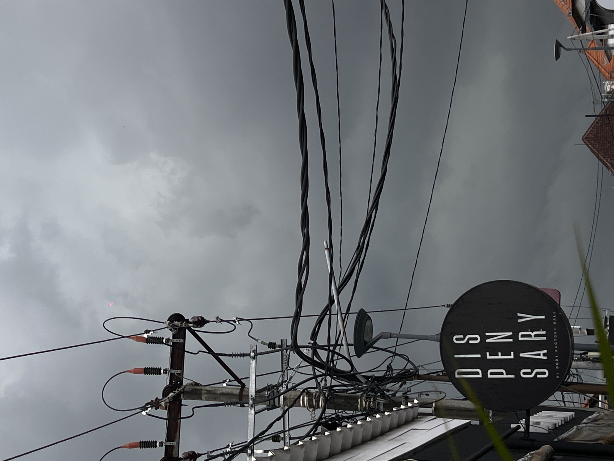 The image shows a view of a cluttered urban scene in North Kuta, Indonesia. Overcast sky with pale gray clouds fills the upper portion of the frame. A dense network of electrical wires dominates the central area, creating a tangled web against the sky. In the foreground, a utility pole with insulators is visible, supporting some of the wires. To the right, a round sign reads "DISPENSARY BY BODY FACTORY BALI". The edges of buildings and rooftops can be seen beneath the wires. Some green plants are in the very foreground, partially obscuring the view.