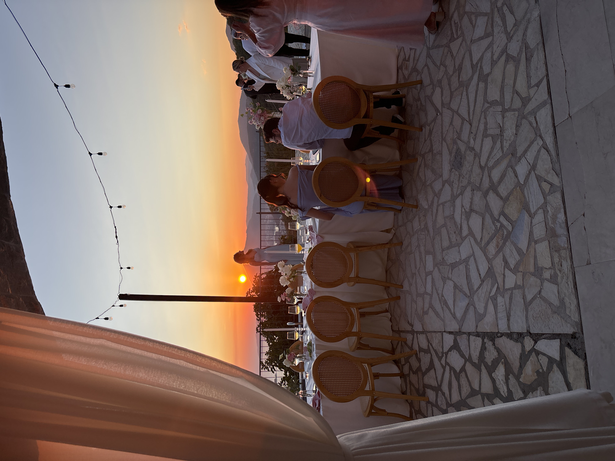 A group of people are gathered at an outdoor dinner party overlooking a sunset and a body of water. The scene appears to be in a Mediterranean coastal region, possibly in a villa or a high-end resort. A long table is set with white tablecloths, floral centerpieces, candles, and wine glasses, indicating a formal occasion like a wedding reception or a celebratory dinner.
There are approximately ten to twelve individuals visible, dressed in semi-formal attire. Several people are seated at the table, engaged in conversation and dining, while others stand and socialize. One woman in a light blue dress is standing near the table, facing away from the camera. Another woman, dressed in white, is taking a photo with her phone. The atmosphere is warm and celebratory, with the golden hues of the sunset casting a soft glow over the scene.
The setting is a paved patio with irregular stone tiles. In the foreground, a large white curtain is draped, framing the left side of the image. In the background, mountains and a distant city can be seen. String lights are hung across the patio, adding to the ambiance. The overall impression is one of a picturesque and intimate gathering, enjoying a beautiful evening.