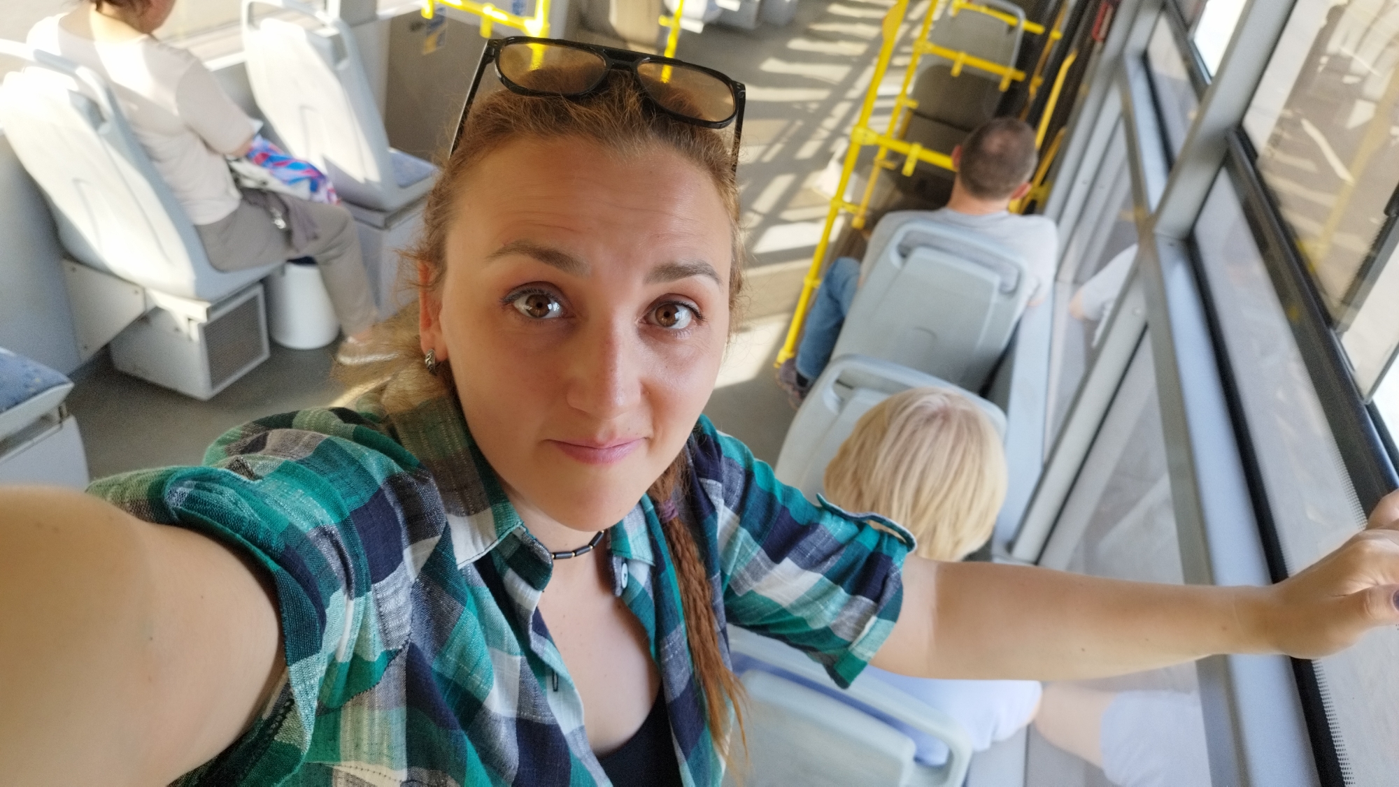 A woman with auburn hair and brown eyes, wearing a green and blue plaid shirt and sunglasses on her head, is taking a selfie on what appears to be a public transport bus. She is seated in the front and the camera is angled upwards. Behind her, other passengers are visible, including a person in a light-colored shirt sitting on the left and a person with blonde hair sitting on the right. Yellow grab bars are present on the right side of the bus, suggesting it is a public bus. The seats are white. The lighting suggests it is daytime. The general setting is consistent with interior public transportation.
