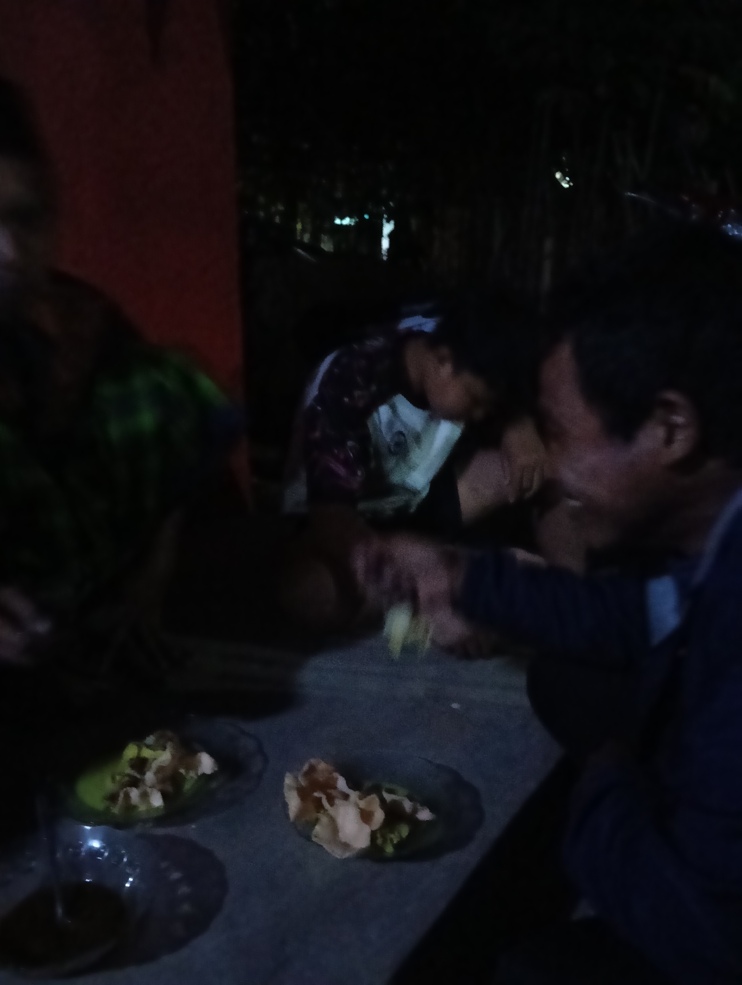 In a dimly lit outdoor setting, possibly a residential yard in Tasikmalaya, Indonesia, several individuals are gathered. The primary focus is on two men in the foreground, seemingly engaged in conversation or a shared activity. One man, on the right, is looking down and to his left, with his mouth slightly open, and his right hand is extended forward, holding a yellow object that appears to be food. The other man, to his left and slightly behind, is also looking down and appears to be reaching for something or interacting with the food. To the left of these two men, another person is partially visible, wearing a green and black patterned garment. In the background, the scene is very dark, with some indistinct shapes suggesting trees or foliage, and possibly some distant lights. On the ground in front of the men are two small, clear plates containing what looks like food, possibly rice crackers or similar snacks, and some green elements. The overall impression is of a casual gathering, perhaps a meal or a social interaction, taking place at night.