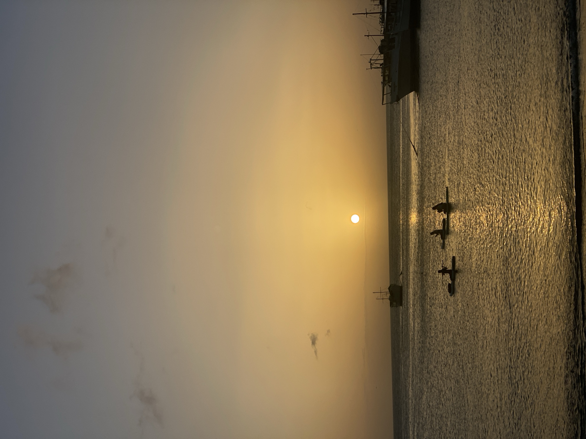 The image depicts a sunset scene over the waters of Juan Griego, Venezuela. In the foreground, the ocean reflects the warm hues of the setting sun, with gentle waves lapping at a dark shoreline. Three individuals are visible on paddleboards in the mid-ground, silhouetted against the water. Further out in the water, a small, traditional sailing vessel is positioned to the left, also silhouetted. To the right, two larger boats are docked, their structures clearly defined against the darkening sky. The sky itself transitions from a pale blue at the top to a rich golden orange near the horizon, with a few faint clouds scattered throughout. The overall atmosphere is tranquil and serene, characteristic of a coastal sunset.