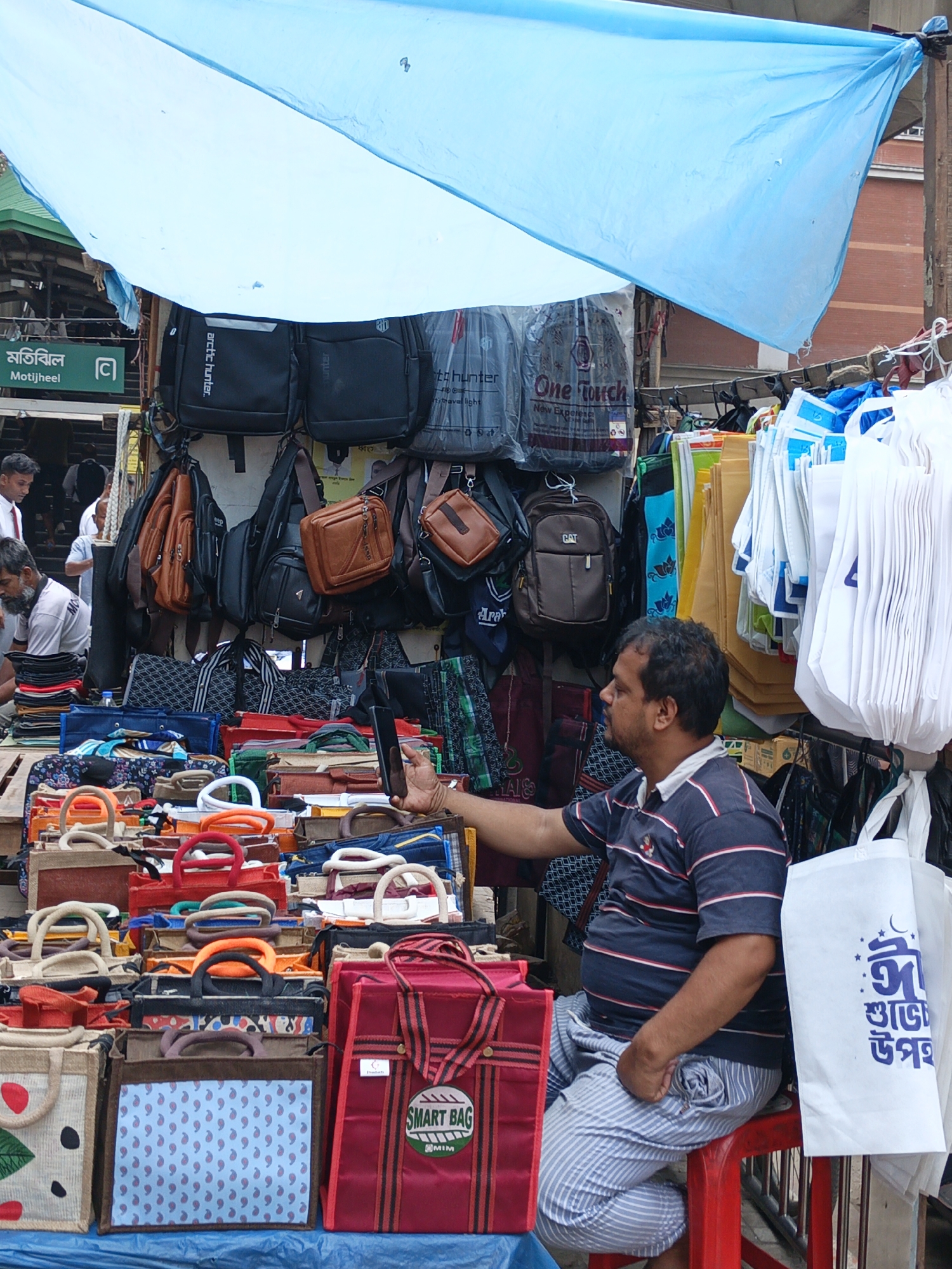 The image shows a vendor sitting on a red plastic stool at an outdoor market in Dhaka, Bangladesh. He is holding a black smartphone with his right hand. The vendor is a middle-aged man with dark hair, wearing a blue and white striped polo shirt and blue and white striped pants. The man is seated in front of a large display of bags. The bags are made from a variety of materials and come in different colors and sizes. The bags are organized in rows, and there are many different styles to choose from. The bags are under a blue tarp for shade. To the left of the vendor, customers can be seen. A street sign that says "Motijheel" can be seen in the background.