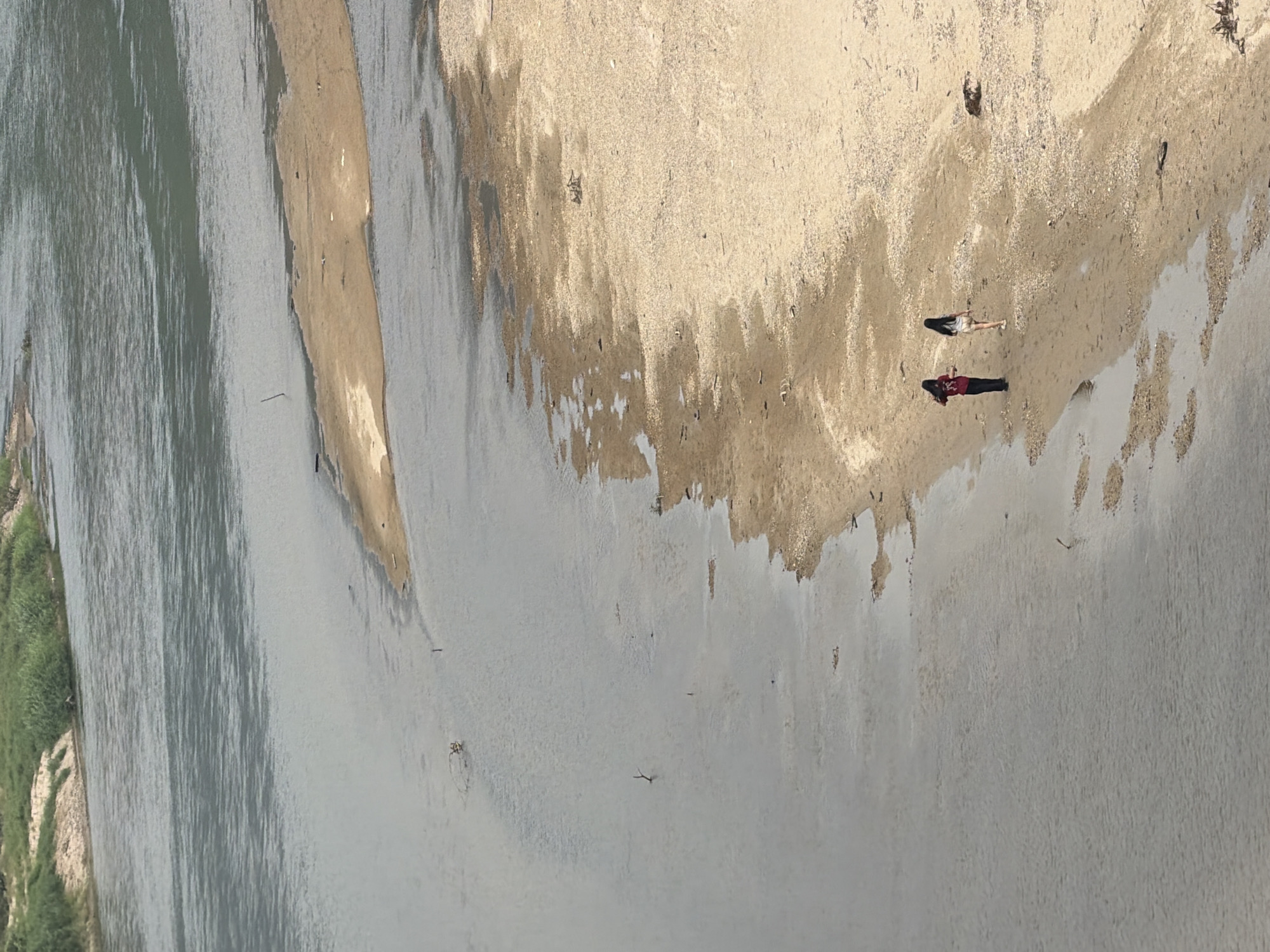Aerial view of a river meeting a sandy bank. Two people are visible on the bank, one wearing a red shirt and the other a light-colored outfit. The environment is characterized by sand, water, and some vegetation along the river's edge. Location is speculated to be in the city of Đà Nẵng, Vietnam.