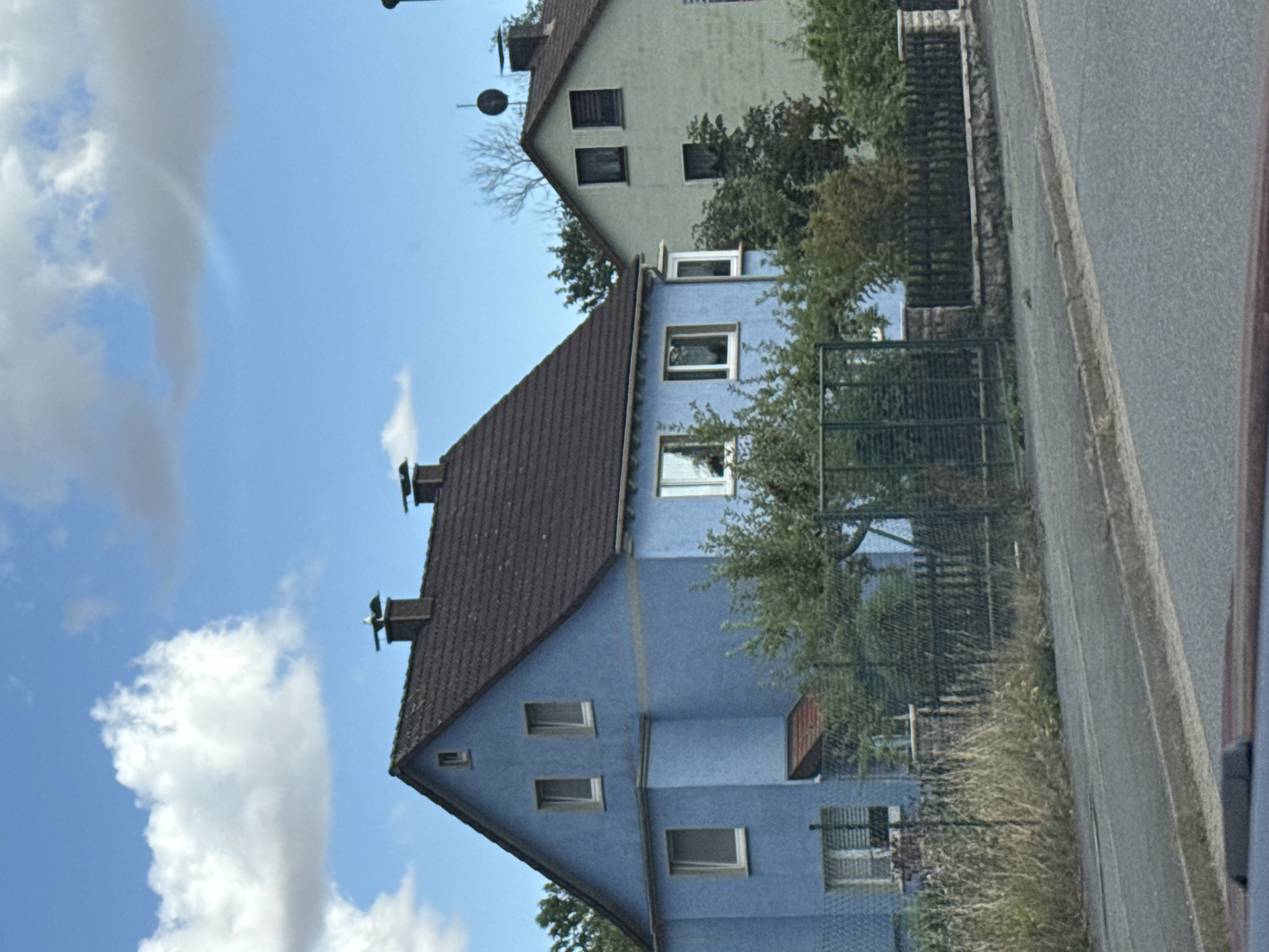 The image captures a residential street scene in Bad Sachsa, Germany. The focal point is a vibrant blue two-story house with a dark tiled roof. This house features several windows of varying sizes and a prominent dormer window on the upper floor. Adjacent to the blue house is a lighter-colored, possibly pale green, two-story house also with a tiled roof. Both houses appear to be residential dwellings.
The foreground of the image shows a paved road, a sidewalk, and a low stone wall topped with a dark wooden fence. Beyond the fence, overgrown dry grass and green foliage are visible, suggesting a somewhat wild or unkempt area. The background is dominated by a bright blue sky with scattered white and gray clouds, indicating fair weather. Trees with green and some autumnal foliage are present, adding to the natural setting. Chimneys with decorative bird figures are visible on the roof of the blue house. The overall impression is one of a quiet, suburban or semi-rural environment.