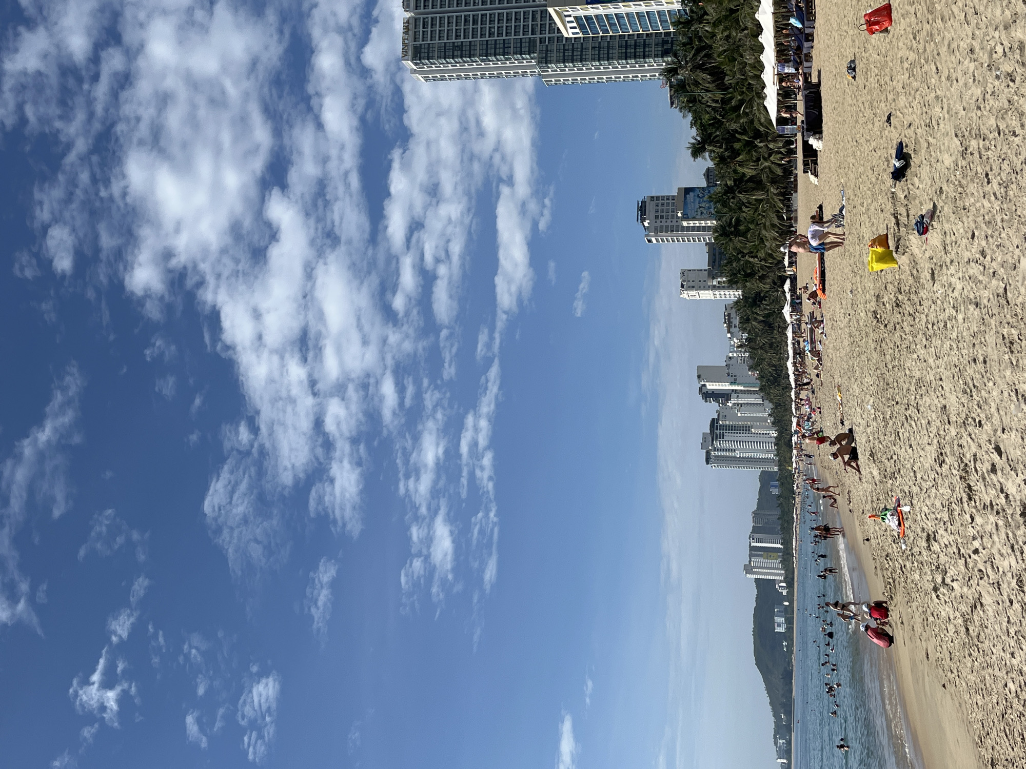The image depicts a crowded beach in Nha Trang, Vietnam, on a sunny day. The foreground is dominated by sand, with several beachgoers scattered across it. Some individuals are sunbathing on towels, while others are engaged in activities near the water's edge. A few beach chairs and umbrellas are visible, suggesting a resort area.
In the mid-ground, the ocean is populated with numerous swimmers and people wading in the shallow waters. The shoreline is bustling with activity.
The background reveals a cityscape with several tall, modern buildings, indicating an urban coastal environment. Lush green palm trees line the edge of the beach, contrasting with the man-made structures. A hilly landscape can be seen further in the distance. The sky is a clear blue with scattered white clouds. The overall scene conveys a lively atmosphere of a popular tourist destination.