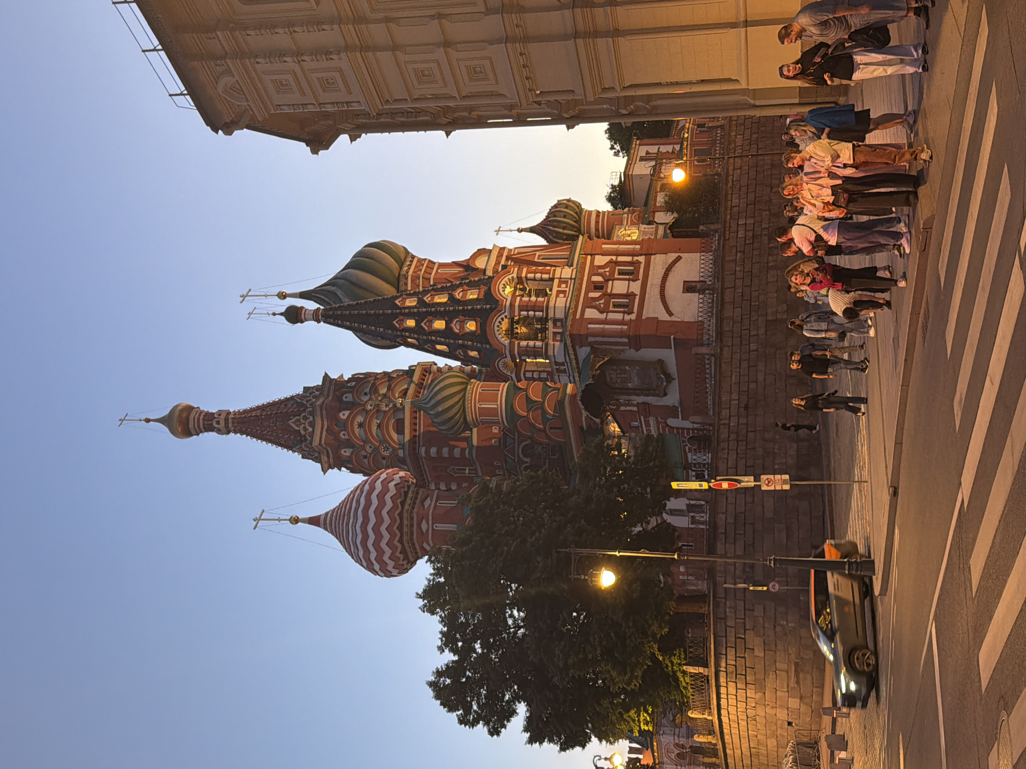 This evening scene in Moscow, Russia, prominently features the iconic St. Basil's Cathedral under a clear, light blue sky, indicating dusk or early evening. The cathedral, standing on the left side of the frame, displays its vibrant red-brick structure and characteristic multi-colored onion domes, including one with distinctive white and red stripes and others in shades of green, gold, and blue. Several spires are topped with golden orbs and Orthodox crosses. Lights illuminate the cathedral's windows, emphasizing its intricate details, and a large, dark green tree partially obscures its lower section.
On the right, a large group of approximately fifteen to twenty people is gathered on a zebra crossing, likely waiting to cross the street or admiring the landmark. They are casually dressed. Further down the street, more people are visible, and a car with illuminated headlights can be seen on the far right.
The street itself is paved with white zebra stripes, and a bright yellow streetlight near the tree casts a warm glow. Other buildings with classical architectural elements, such as patterned facades, are visible on the far left, towering above the cathedral. A stone wall lines the street, and various street furniture, including poles holding traffic signs, are present. Two red circular traffic signs are identifiable: one with a white horizontal bar (likely "No Entry") and another with a diagonal bar (possibly "No Stopping" or "No Parking"), though any specific text is not discernible. The overall atmosphere is that of a lively urban evening at a historic site.