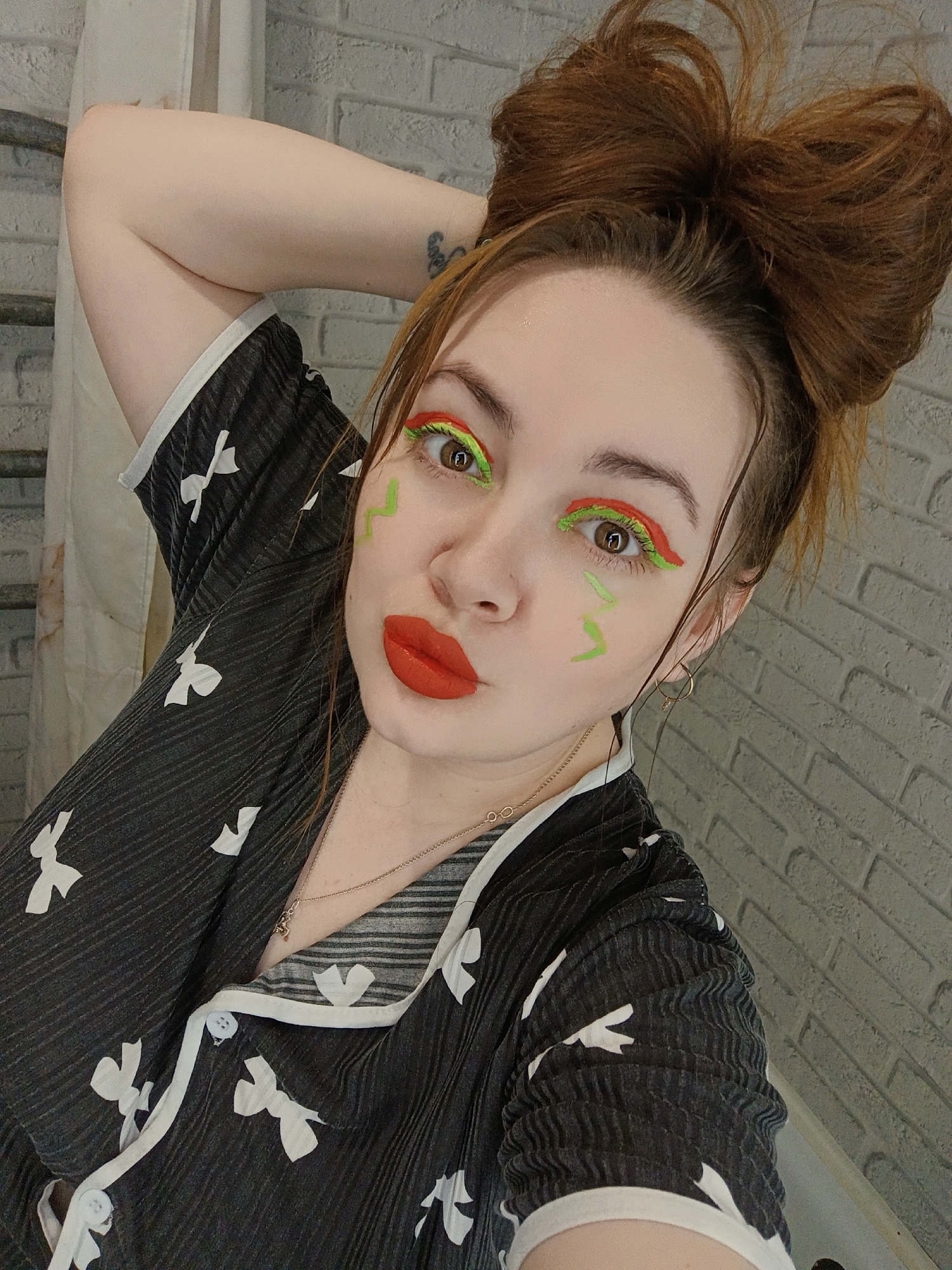 A young woman with reddish-brown hair, styled in a voluminous messy bun, poses for a close-up selfie indoors in Chaikovsky, Russia. She has fair skin, and her left arm is raised behind her head, revealing a small, indistinct tattoo on her upper arm.
She is wearing a black, pleated top or pajama shirt adorned with numerous white bow or ribbon patterns. The garment features a white collar, white trim, and white buttons down the front placket. Her makeup is a notable detail, characterized by bright red lipstick on her pursed lips, and bold neon green and orange eyeliner and eyeshadow around her eyes, creating a graphic, layered look. Neon green squiggle designs are also painted on her cheeks below her eyes. She accessorizes with small gold hoop earrings and a delicate gold-colored necklace with a tiny charm.
The background shows a light grey wall with a brick-like pattern. To the left, a portion of a white curtain or shower curtain and a metal fixture are visible. The woman's expression, combined with her vibrant makeup, suggests a playful and expressive mood. There is no clearly legible text visible in the image.