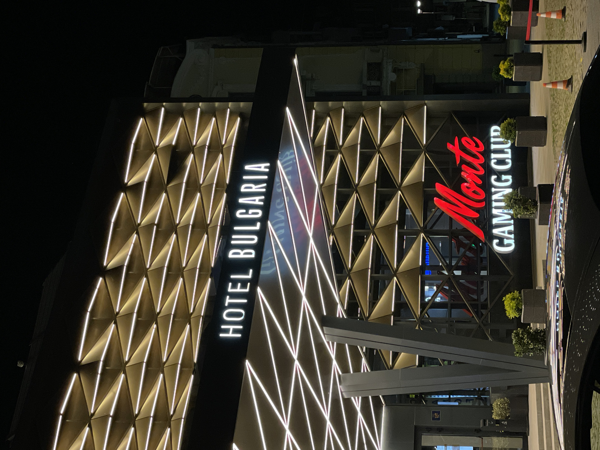 The photograph shows the facade of "Hotel Bulgaria" and "Monte Gaming Club" at night in Burgas, Bulgaria. The hotel has a geometrically patterned facade with illuminated white lines. The casino's name is in red, adjacent to the words "GAMING CLUB" in white. Planters with greenery line the entrance. A car is partially visible on the right. Cones are placed along the entry lane.