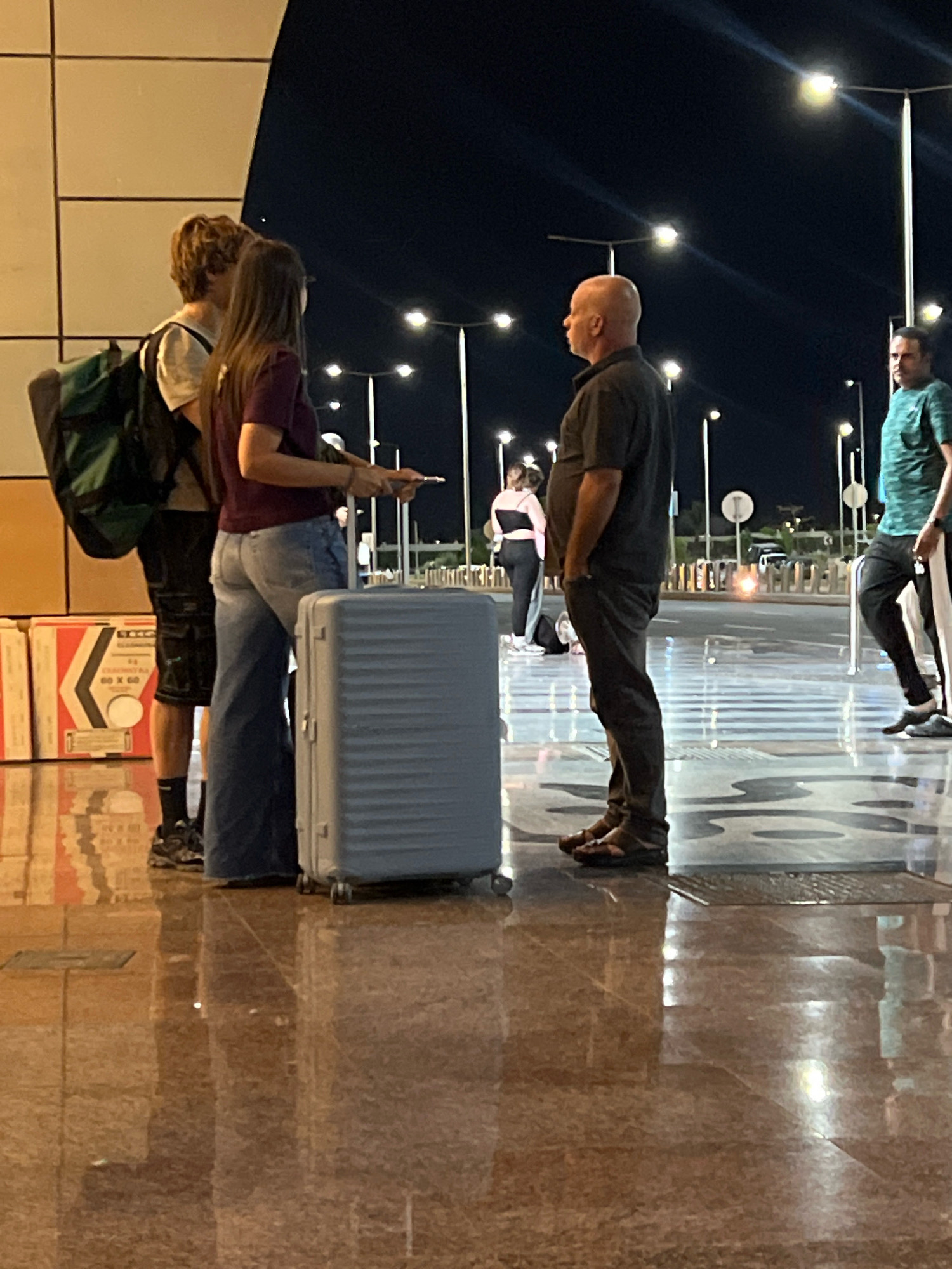 At night, in an outdoor area in Sharm El Sheikh, Egypt, several individuals are present. A young man with blond, tousled hair stands facing left, wearing a white t-shirt and dark shorts, with a green backpack on his left shoulder. Next to him, a young woman with long brown hair, wearing a maroon t-shirt and light blue flared jeans, faces towards the viewer's right. She holds a smartphone in her right hand. Between them and slightly in front is a light blue, rectangular suitcase on wheels. To the right of the couple, a bald man with dark skin, dressed in a dark gray polo shirt and dark pants, stands facing left, with his hands in his pockets. Further to the right and slightly behind, another man with dark skin, wearing a teal striped t-shirt and dark pants, faces towards the viewer's left. In the background, streetlights illuminate the scene, and a dark sky is visible. Reflective flooring is present in the foreground. Boxes are stacked to the left of the young man.
