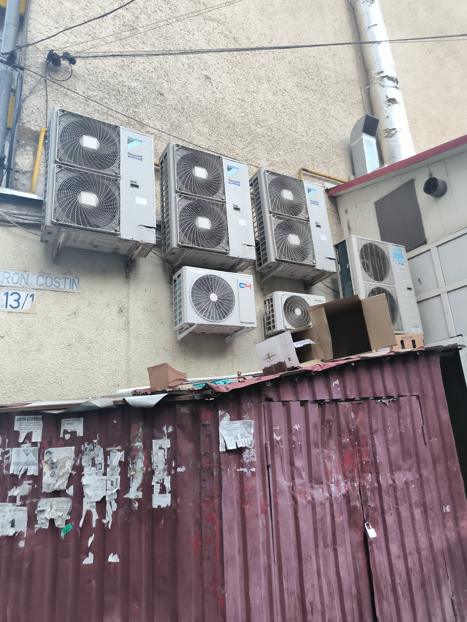 The image depicts an exterior view of a building in Chișinău, Moldova, characterized by a textured beige wall and a corrugated metal structure below. Mounted on the wall are multiple outdoor air conditioning units, several of which are Daikin brand. Some of these units are stacked, with two pairs of Daikin units prominently displayed. Below these, additional air conditioning units from brands like CH and NeoClima are visible. A street sign reads "ARON COSTIN 13/1". The corrugated metal structure, painted a dark red, is adorned with numerous torn posters and advertisements. A white padlock secures a vertical seam on this metal structure. The environment appears urban, with electrical wires running along the wall and a large metallic pipe extending vertically in the background.