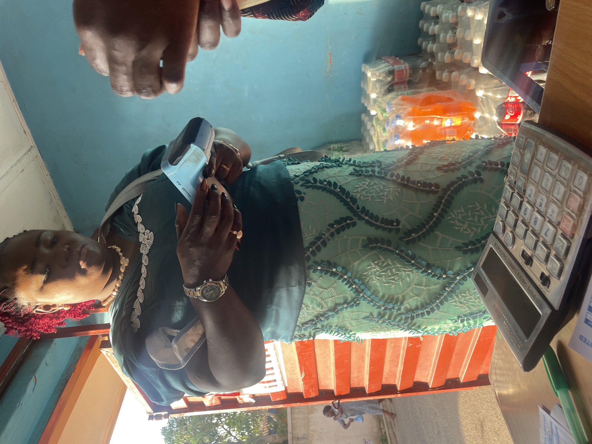The image shows an African woman seated behind a counter inside a shop in Jos, Nigeria. She is wearing a green dress with a floral pattern, a gold necklace, and a watch. In her hands, she holds a small electronic device. A calculator sits on the counter beside her. Behind her are stacks of bottled drinks. Above, a person's hand appears to be reaching towards the counter. Outside, a young girl is walking along the street. The scene suggests a small retail establishment in an urban setting.