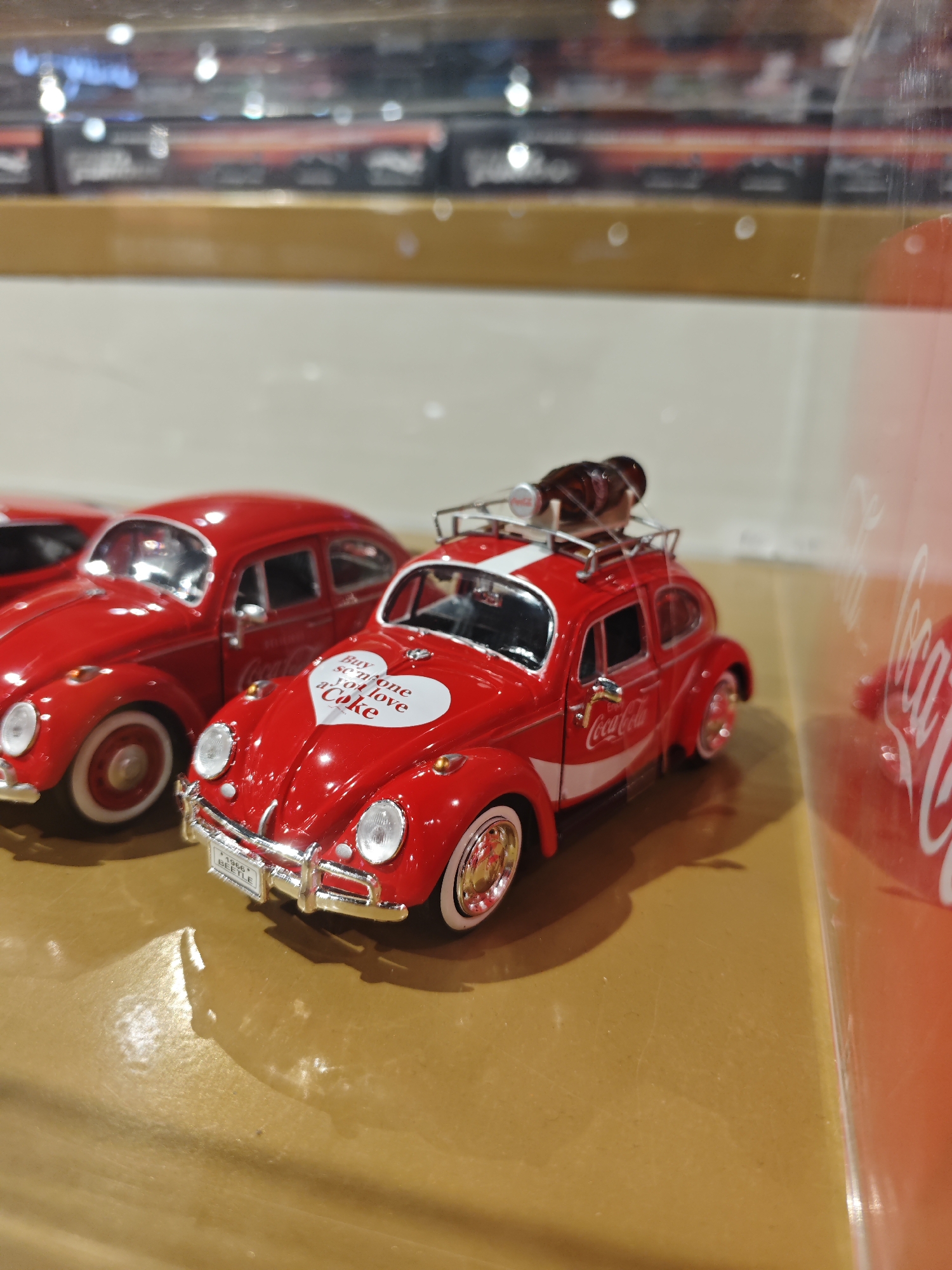 The image shows a close-up of several red toy Volkswagen Beetles with Coca-Cola branding. The Beetles are detailed replicas, with one in the foreground featuring a heart-shaped decal on its hood that reads "Buy someone you love a Coke." This car also has a roof rack with several brown glass Coca-Cola bottles. Another red Beetle is visible to the left, with a similar Coca-Cola design. The models are placed on a reflective, yellowish surface, possibly a table or display. In the background, blurred shelves with more items are visible, suggesting the setting might be a store or display case. The overall lighting is bright, highlighting the glossiness of the toy cars. The environment is indoors and appears to be a retail or display setting in an unknown location in Unknown, Unknown.