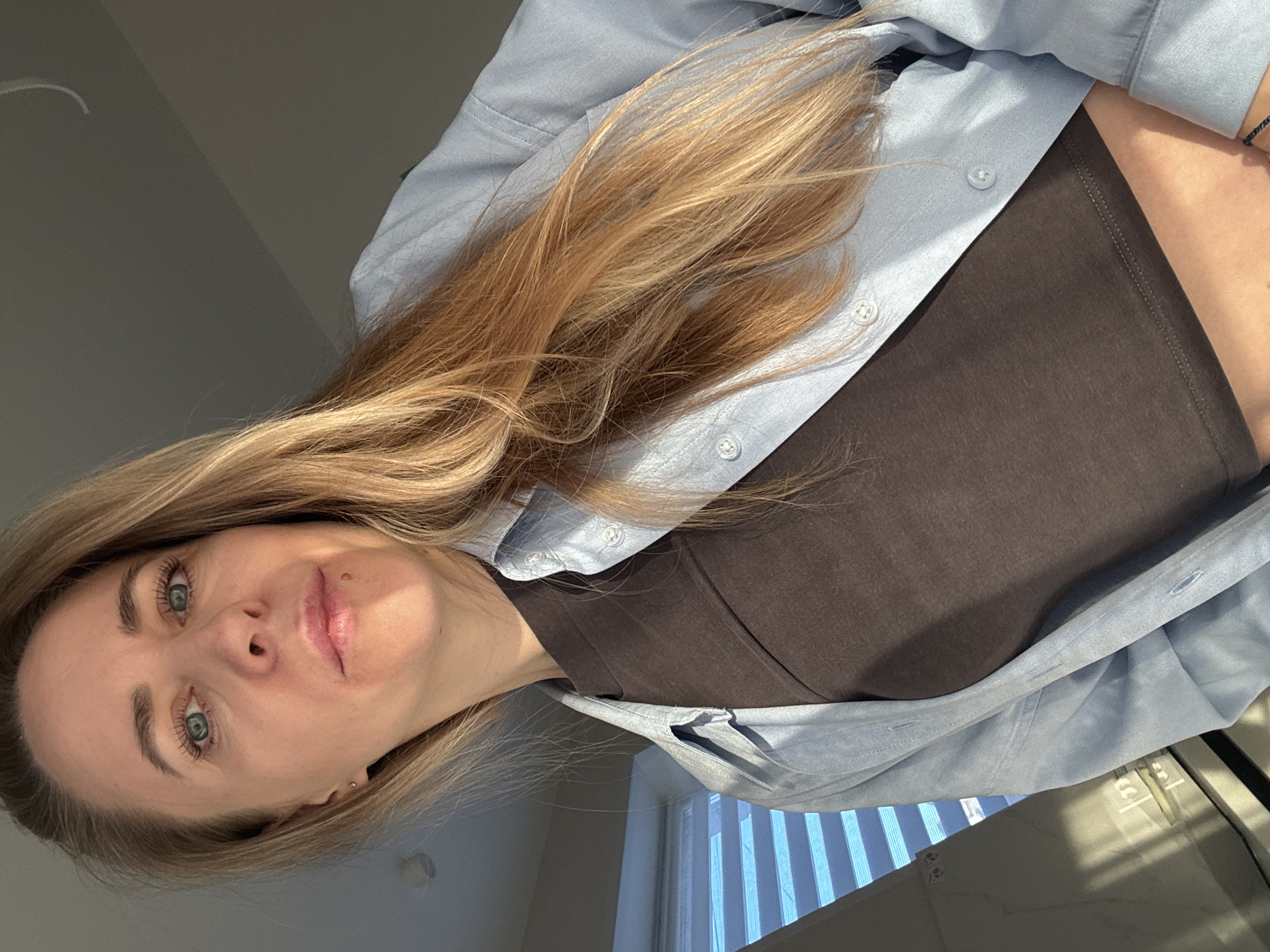 The image, rotated 90 degrees clockwise, presents a close-up indoor selfie of a young adult woman. She has long, flowing blonde hair, light blue-green eyes with mascaraed lashes, and subtly made-up eyebrows. Her lips are naturally colored and full, and a small stud is visible in her left earlobe. She is dressed in a dark brown crop top worn beneath an unbuttoned, light blue long-sleeved collared shirt, possibly chambray. A slim black band or bracelet adorns her right wrist.
The background reveals an indoor setting, likely a room, illuminated by bright natural sunlight entering through a window partially visible on the right. The window features vertical blinds, and the strong sunlight creates distinct highlights on the woman's hair and face, indicating it is daytime. Shadows suggest direct sun exposure. The woman is looking directly into the camera, implying she is taking her own photograph. Her facial expression is neutral and composed. No discernible text is present in the image. This photograph was captured in Chaikovsky, Russia.