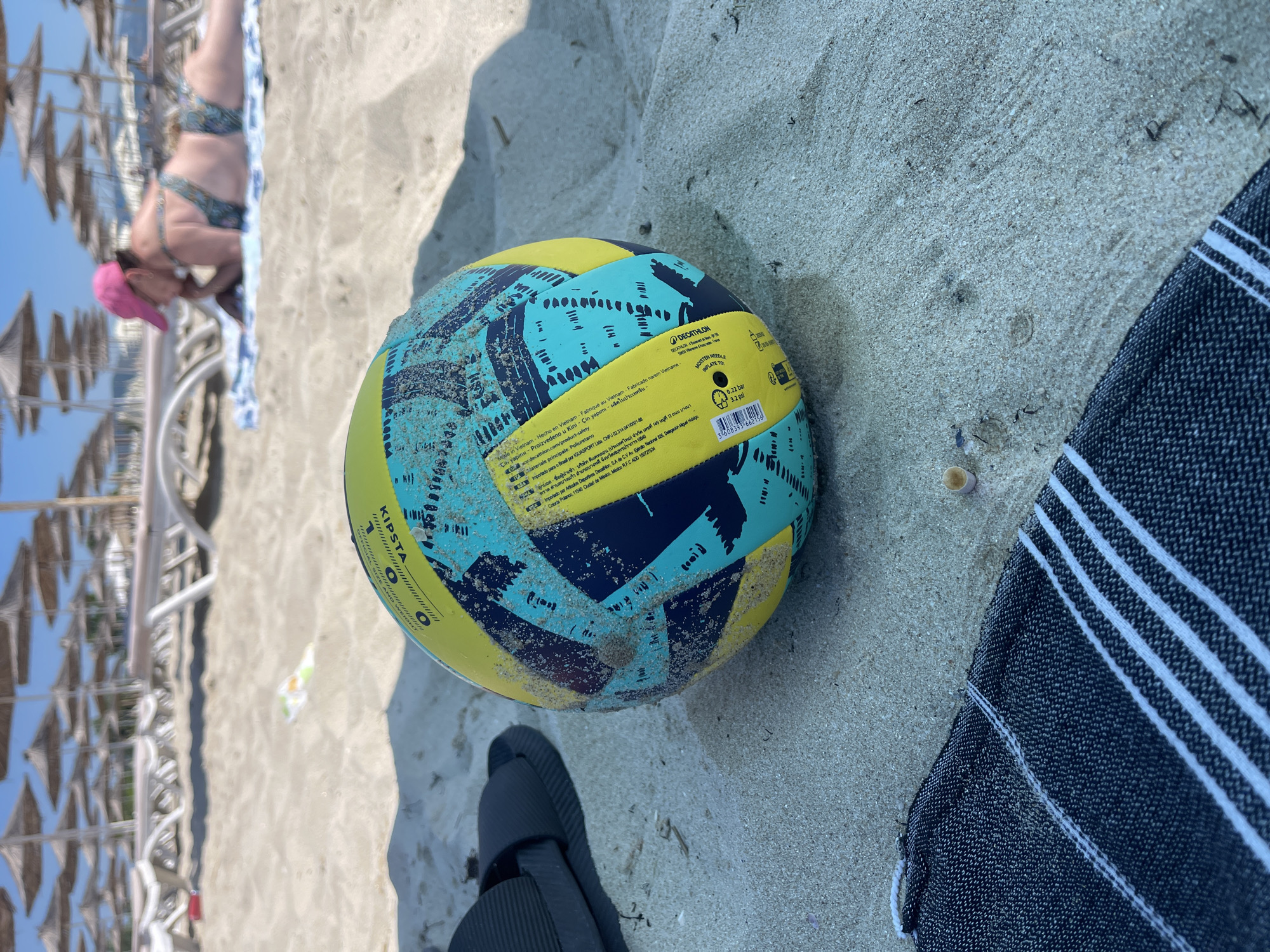 The image shows a volleyball lying on a sandy beach. In the background, beach umbrellas and lounge chairs are visible, suggesting a sunny day at the seaside. A person in a bikini and a pink hat is partially visible in the upper left corner, lying on a towel or beach chair. The volleyball is yellow and teal with dark blue markings and is covered in a light dusting of sand. Text is visible on a label attached to the ball, including "KIPSTA," "DECATHLON," and information about its origin and recommended inflation pressure. A dark-colored sandal is also visible in the lower part of the image, and a striped towel is to the right of the volleyball. The scene depicts a relaxed beach setting, likely during daylight hours.