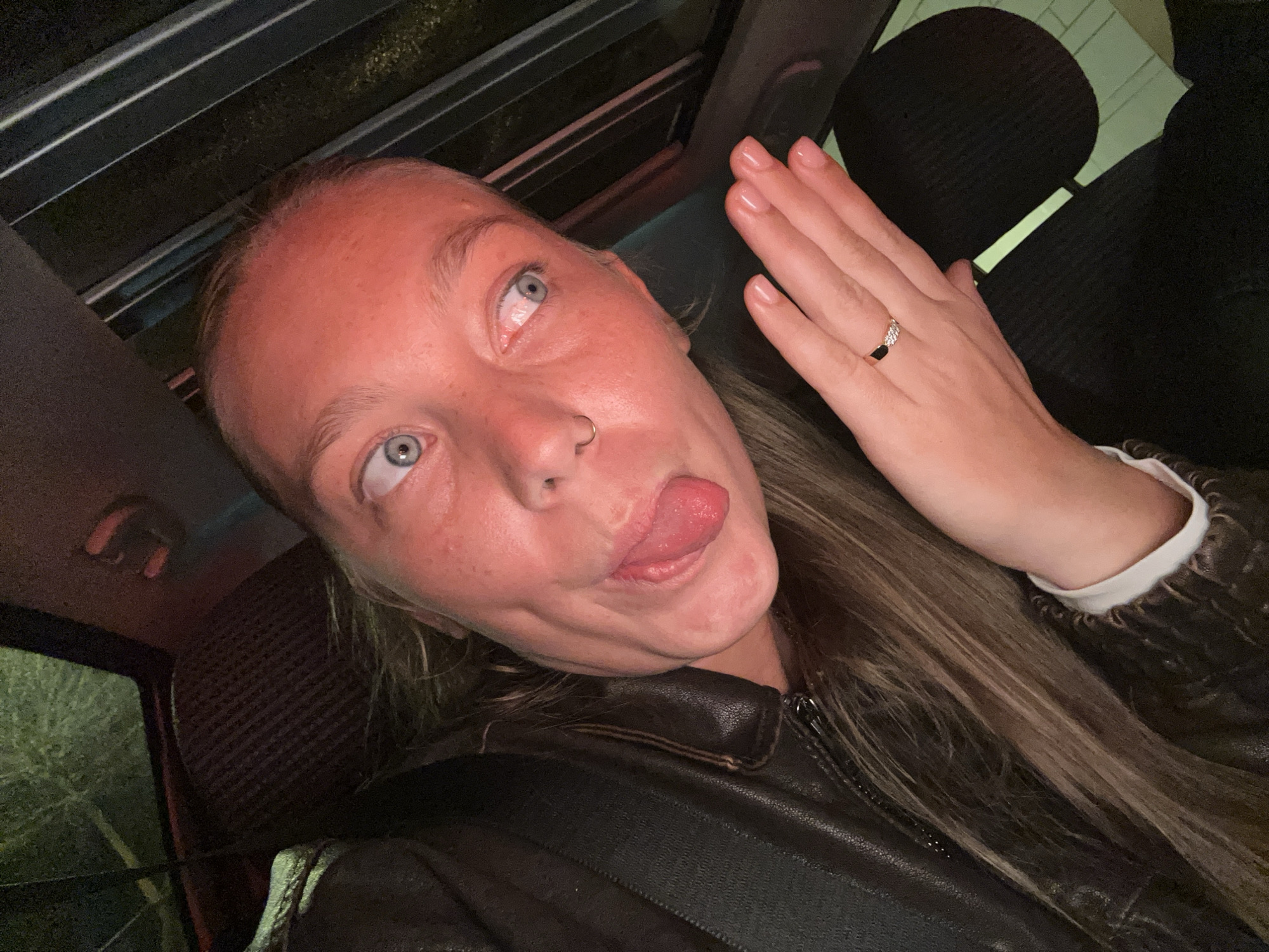 The image shows a woman in what appears to be a car, based on the seat and a glimpse of the car's roof and window. The woman has fair skin, light-colored eyes, and is sticking out her tongue, making a silly face. She has a nose ring in her left nostril. She is wearing a brown leather jacket and a seatbelt. There is a ring on her right ring finger. The lighting is dim, and the background outside the car window is blurry.
I cannot verify the location as Noisy-le-Grand, France, based solely on the image. More context would be required to confirm the geographical location.