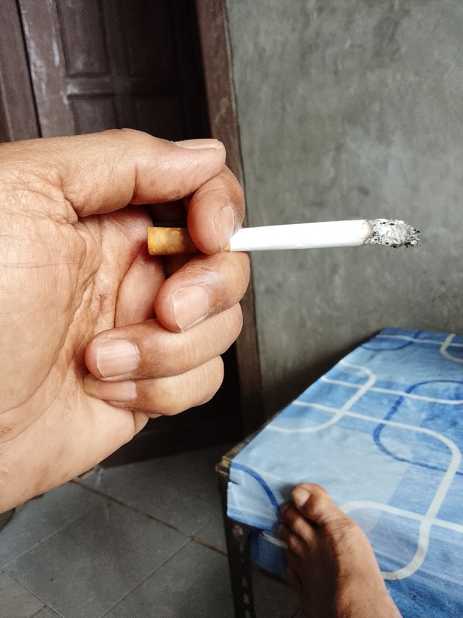 A close-up shot focuses on a hand holding a lit cigarette. The cigarette is white with a filter, and the end is glowing red and covered in ash. The hand, with its skin texture visible, holds the cigarette between the thumb and index finger, with other fingers curled underneath. In the background, out of focus, is a gray textured wall and a partially visible wooden door. To the right, a portion of a surface covered with a blue and white patterned cloth is visible, with a bare foot resting on it. The floor appears to be tiled with dark gray square tiles. The scene has the appearance of an indoor setting, possibly a room.