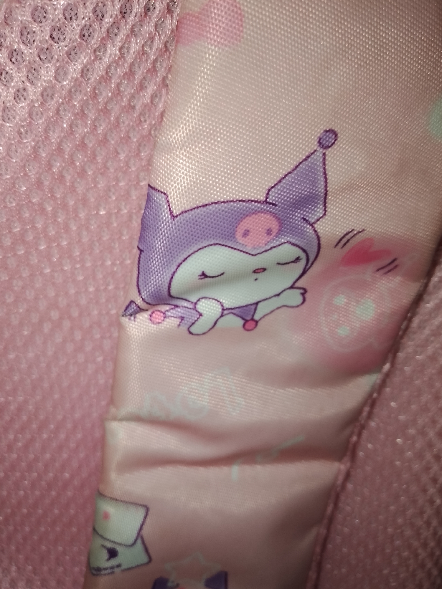 The image shows a close-up of a pink fabric with a cartoon character printed on it. The character appears to be a purple bat-like creature with closed eyes, a pink nose, and small pink ears. It is lying down with its hands up, and there are small pink hearts floating around its head. The fabric is textured and has a slightly shiny appearance. The background is also pink and appears to be a mesh material. There are no discernible landmarks or elements that would place this image in Santa Ana, Venezuela.